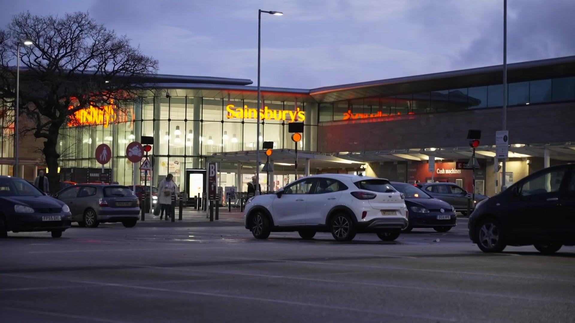 Cars are parked outside the Sainsbury's store on Wilmslow Road in Cheadle. The image is taken at dusk with the inside of the supermarket lit up by artificial lighting. The store's orange logo is prominently displayed in lights.