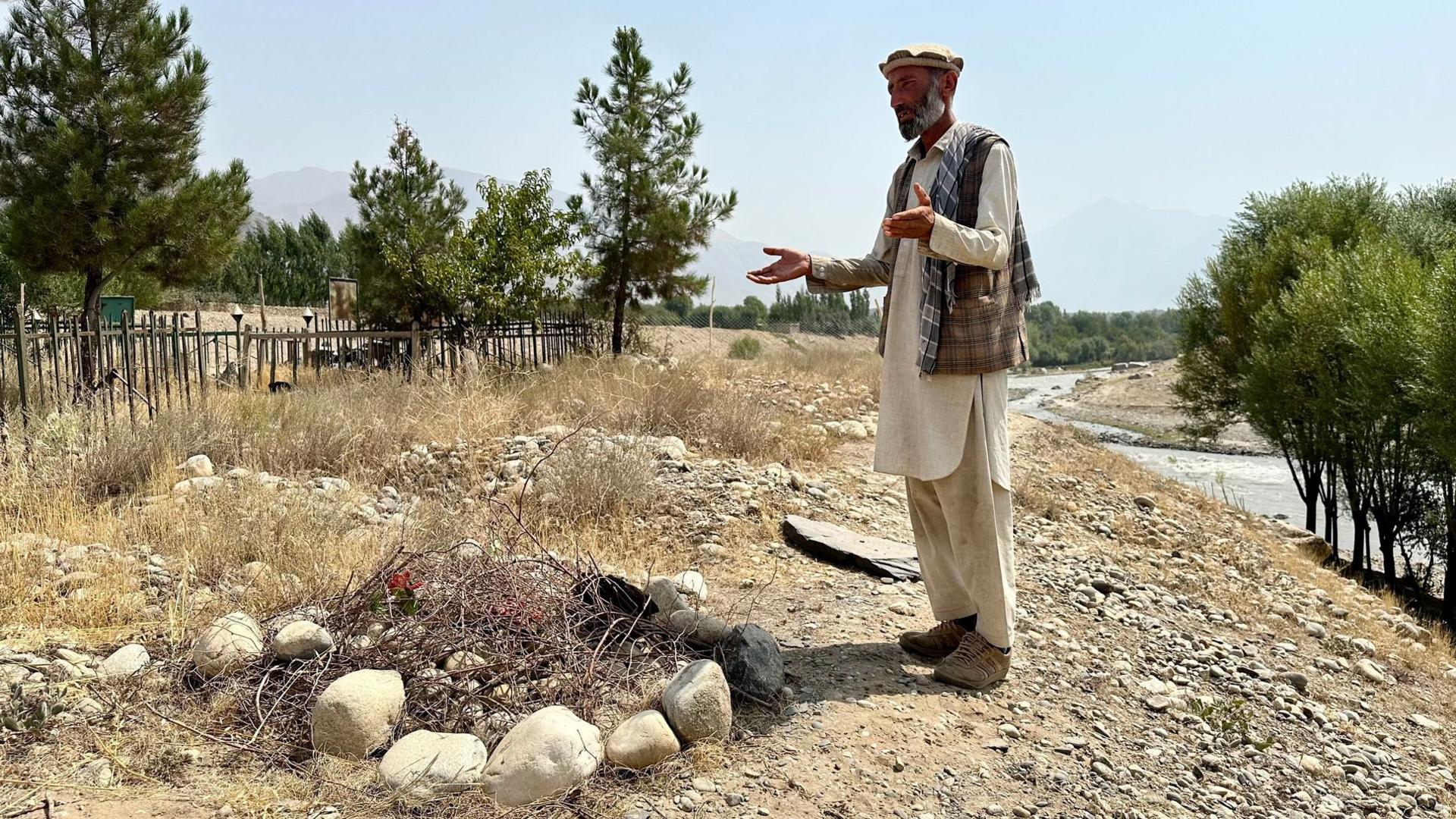 Abdul gestures as he speaks while stood next to the grave of his newborn baby. The grave is marked by a collection of rocks and foliage against a background of trees, river and blue sky in the village of Shesh Pol.