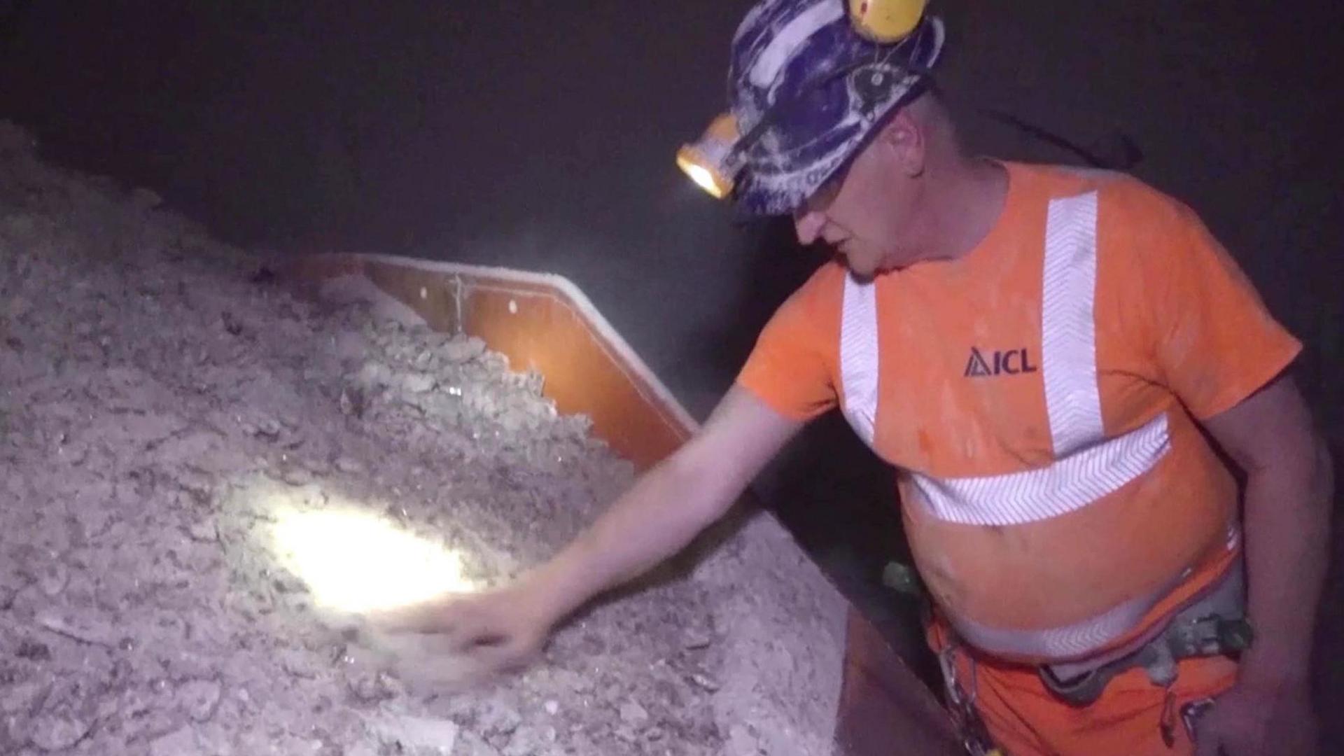 A man working in Boulby Mine. He can be seen touching a pile of rock salt and is wearing orange overalls and hard hat with a torch lamp on top. 