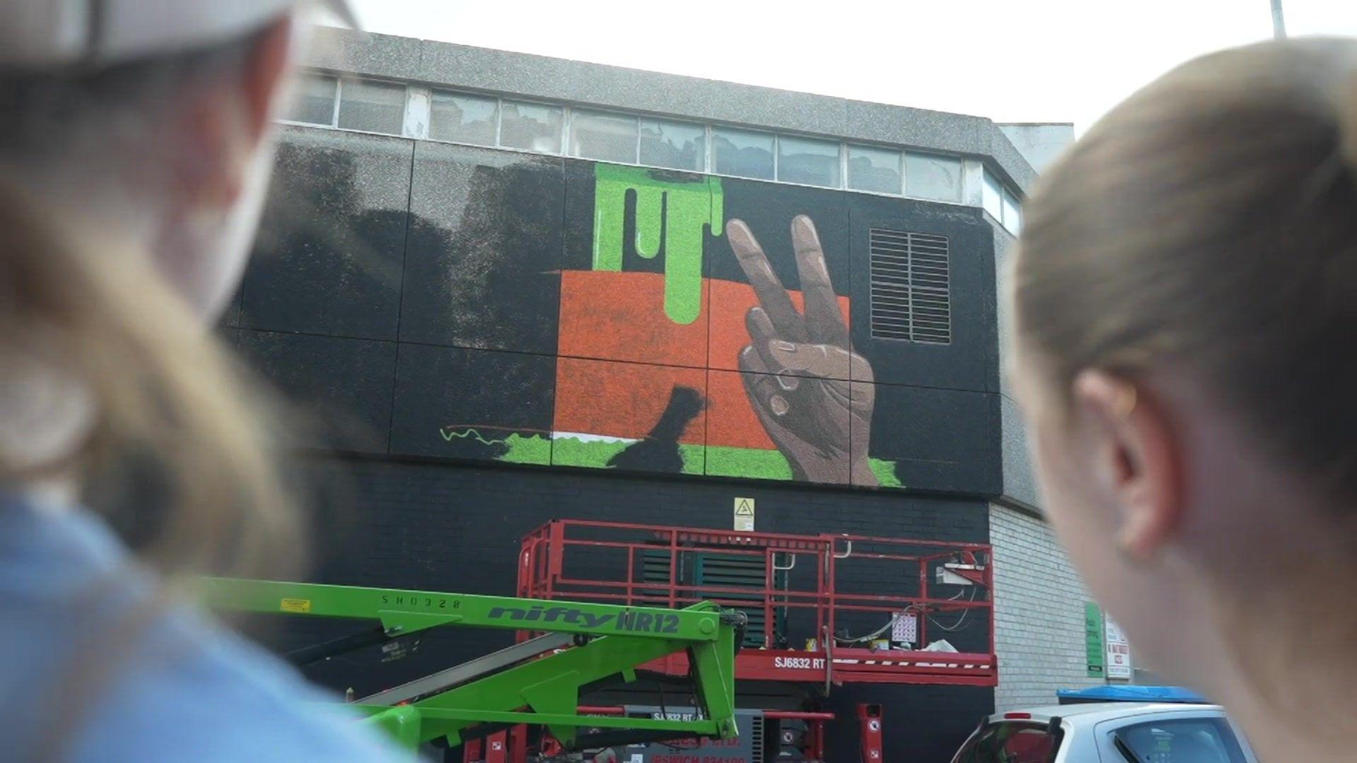 Two artists are pictured looking at a mural that is mid-painting on a building in Ipswich. The mural shows a hand making a peace sign against a black, orange and green background.