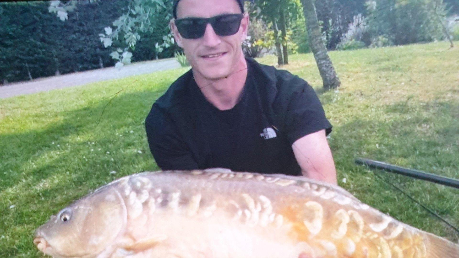 Louis Price with a large fish. He is wearing a black t-shirt and there is grass and trees behind him. 