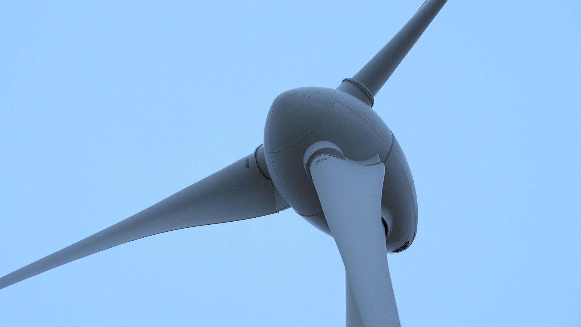 A close up of the centre of a wind turbine. Three white blades are attached to the dome shaped centre which stands on top of a large mast.