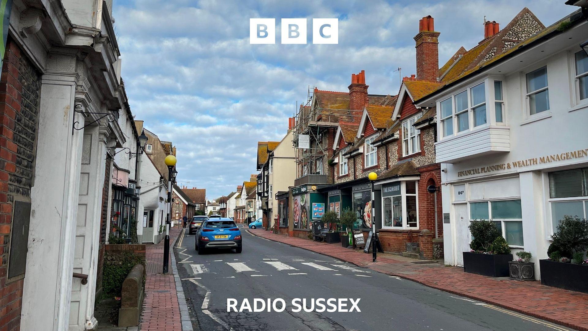 Looking down the road in Rottingdean village centre. Old 2 story buildings line the street on either side and there is a zebra crossing across the road. A blue car drives away from the camera.