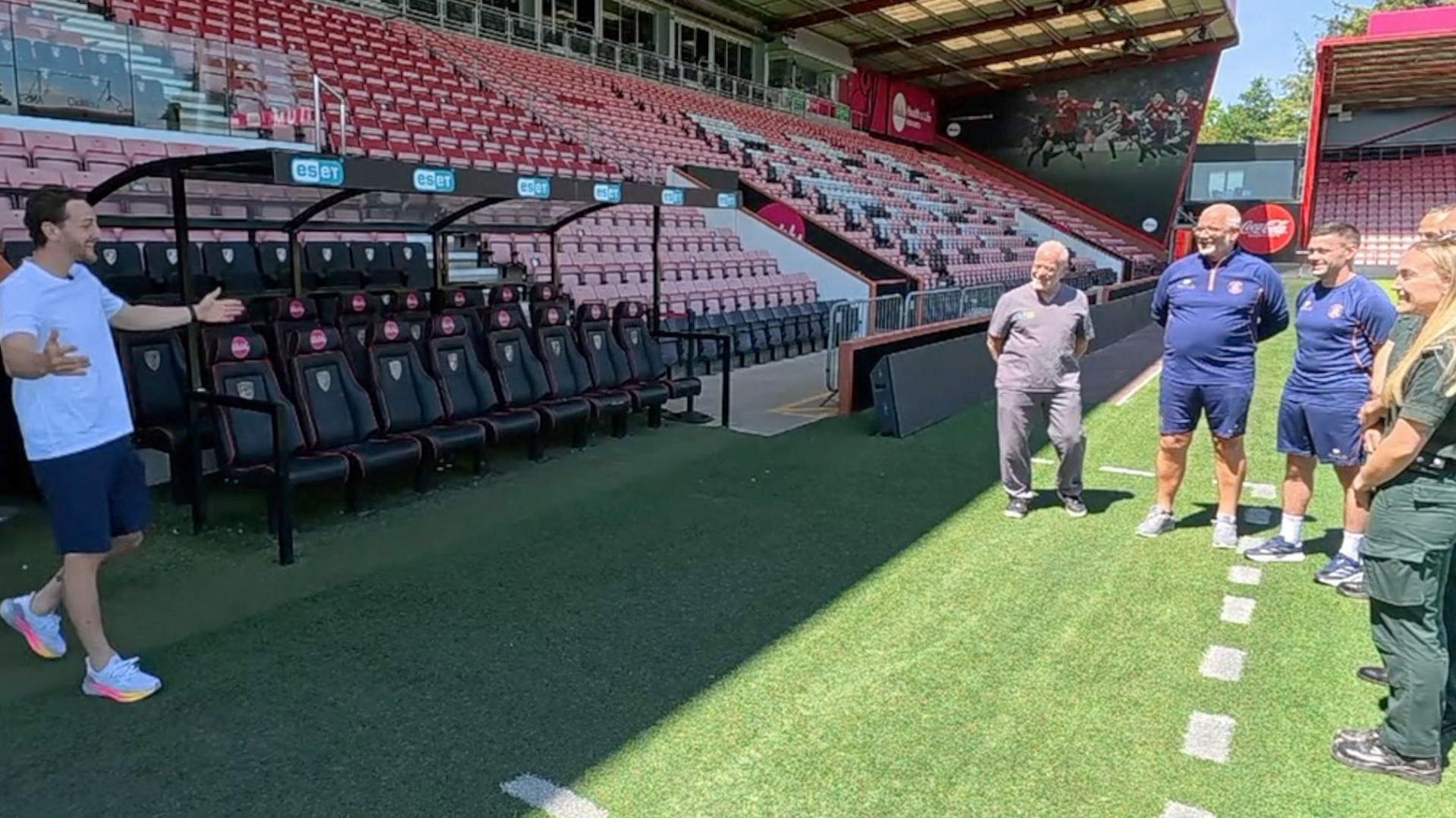 Tom Lockyer is wearing a white T-shirt with blue shorts and white running shoes with pink and orange-trimmed soles. He is walking out on to a football pitch of an empty stadium with his arms outstretched towards two paramedics and three men in football club training gear. They are standing in the sun on the edge of the manager's technical area - signified by a white dotted line painted on the grass - while Tom is in the shade cast by the stand above him