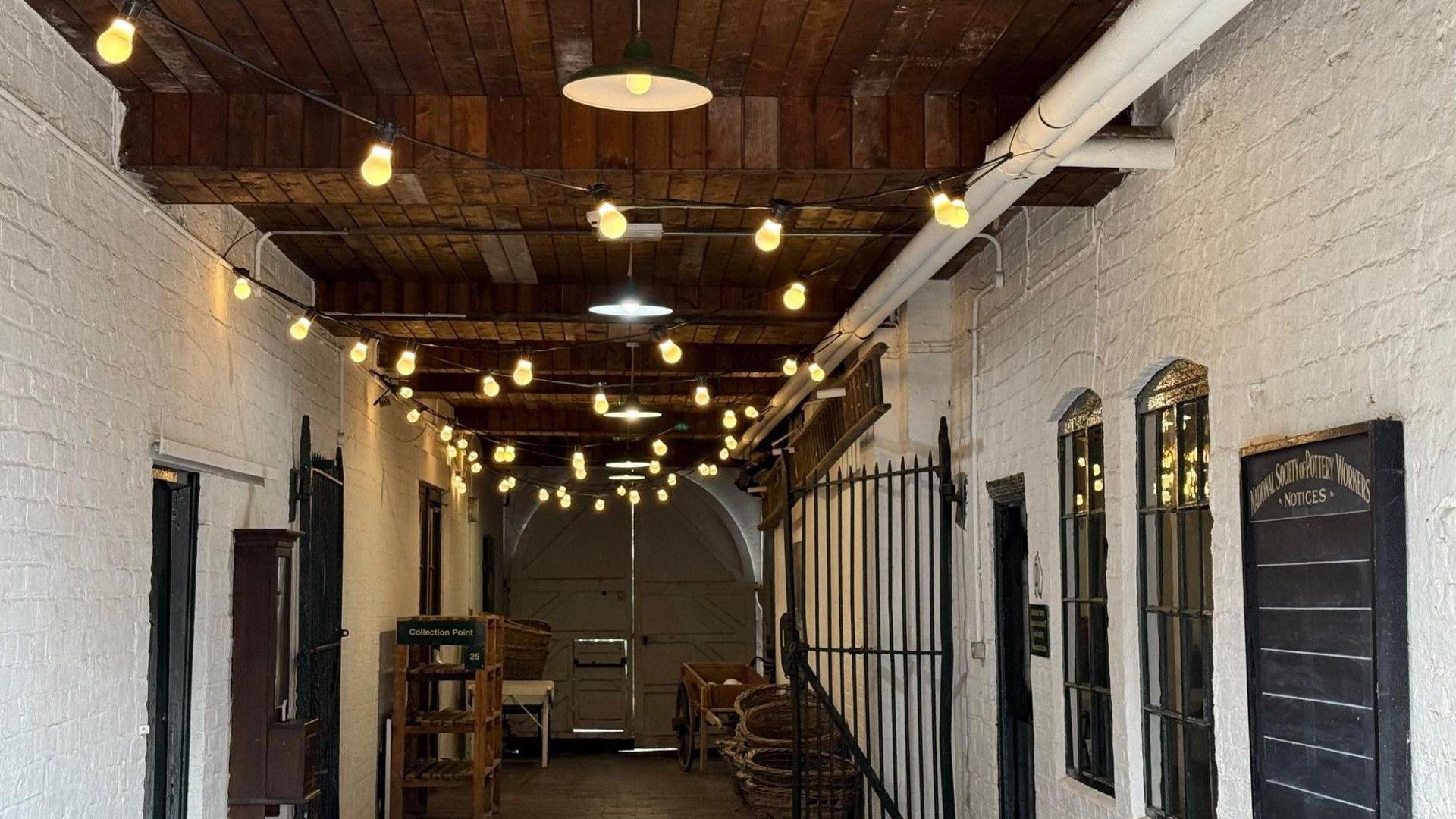 A corridor with festoon lighting strung across, with white-painted bricks and large factory double doors at the end. There are black metal gates attached to the walls.