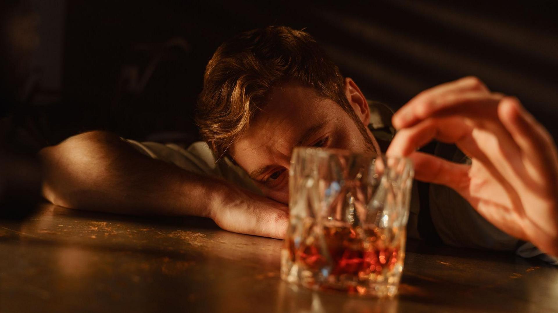 A man with brown hair and a beard leans his head on his arm on a table and gazes into a glass of whisky while running his index finger around the rim of the glass.