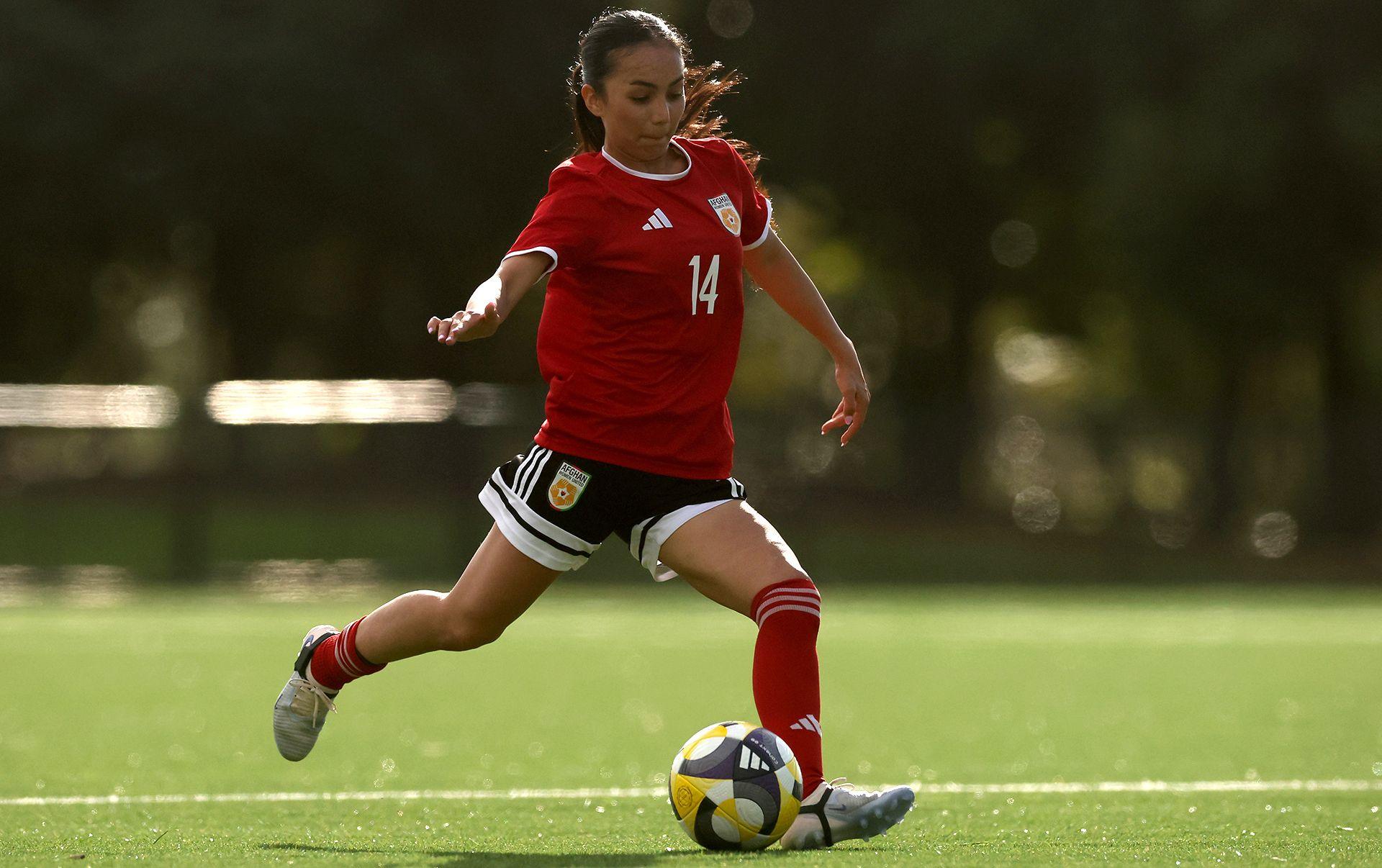 Action changeable  of a pistillate   astir  to footwear   a football. She is wearing a reddish  garment  with the fig   14 connected  it successful  white. She has achromatic  shorts, reddish  socks and achromatic  boots. Her agelong  acheronian  hairsbreadth  is tied successful  a ponytail. 