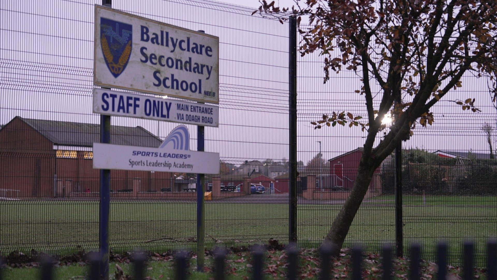 Exterior image of a brown school with a green field in front of it. In the foreground there is a large fence, a tree and signage showing Ballyclare Secondary School entrance