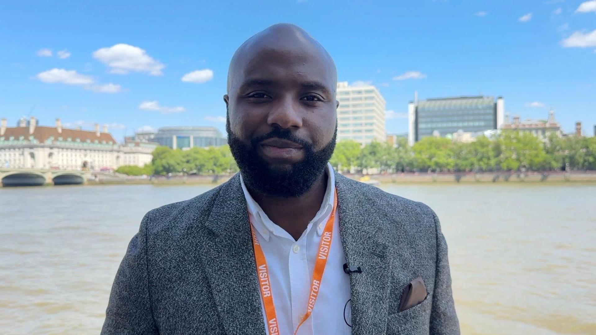 A man with a black beard. He is wearing a suit and standing in front of a big inner city river.