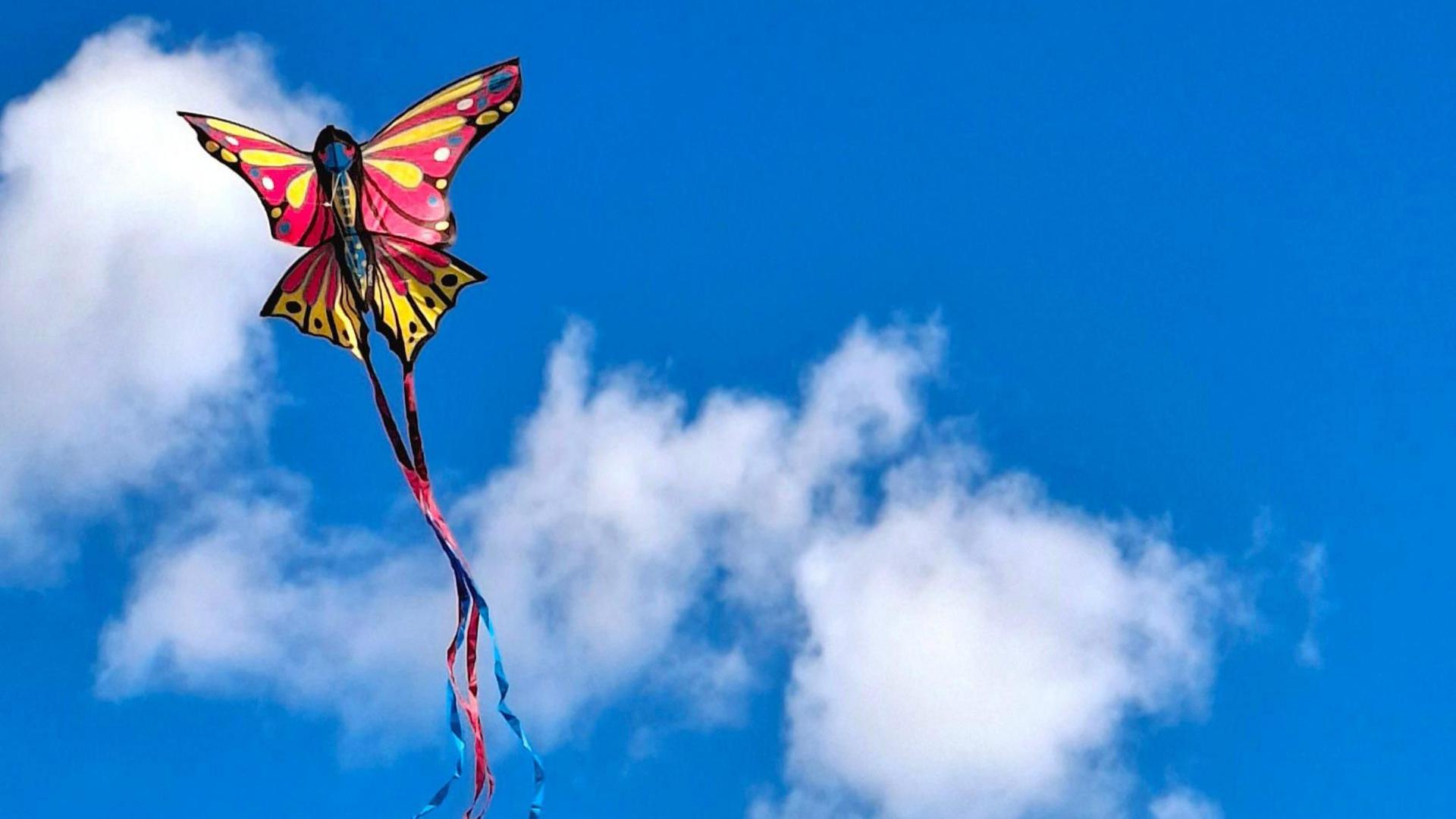 A colourful butterfly kite in the sky over Slough. The kite is the shape of a butterfly with pink and yellow wings with a blue and red tail. It is flying on a sunny day against a bright blue sky with small fluffy white clouds. 