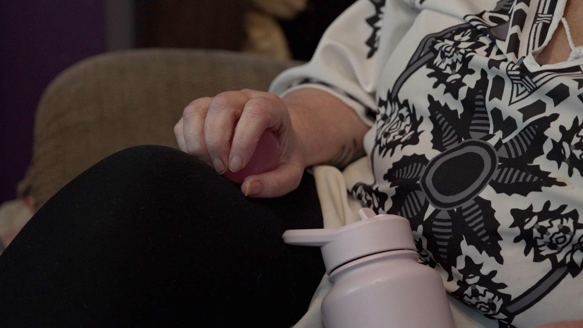 A close-up of Lorraine's hand clutching a squishy stress ball. She is wearing a white top with a black design of a stylised sun or flower. Her pink-purple water bottle is visible in the frame.