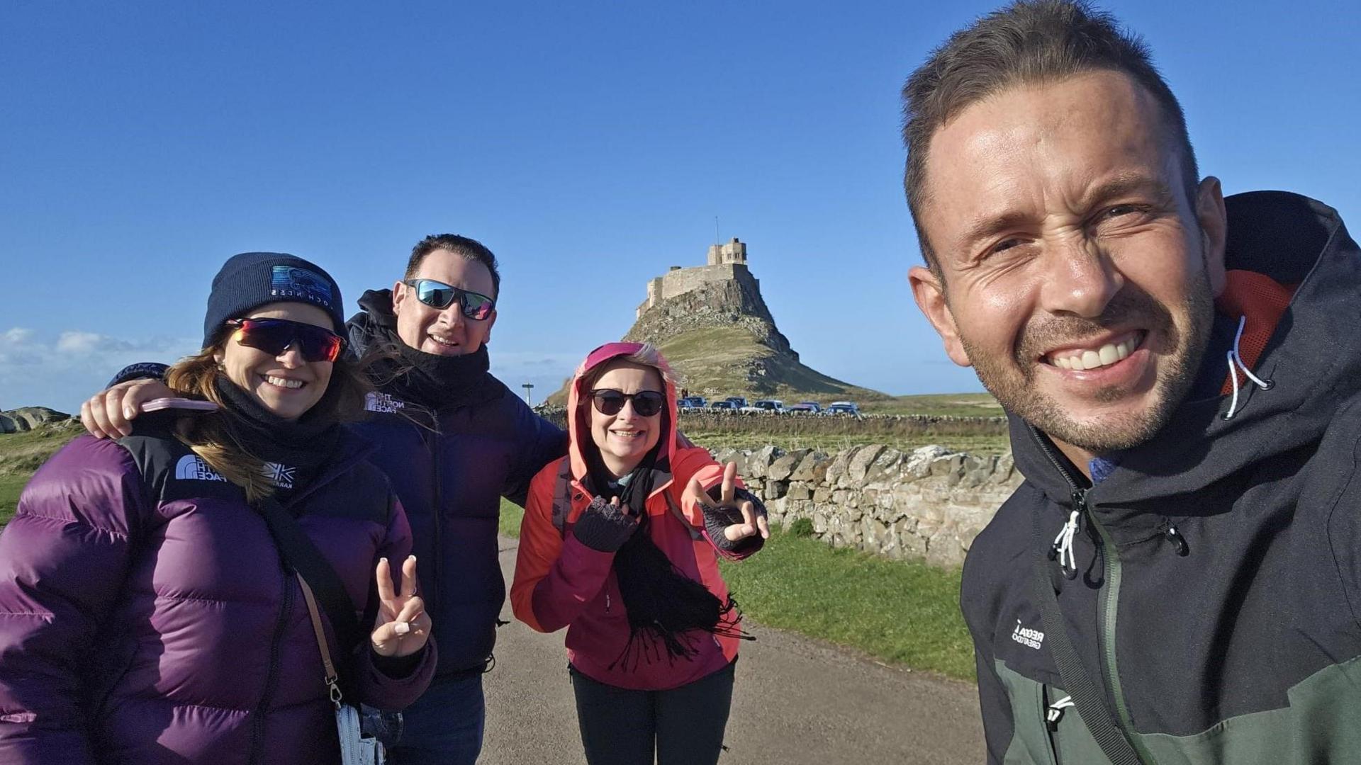 The picture shows Dita and Jesus Sanchez and two friends smiling into the camera with Lindisfarne Castle behind them