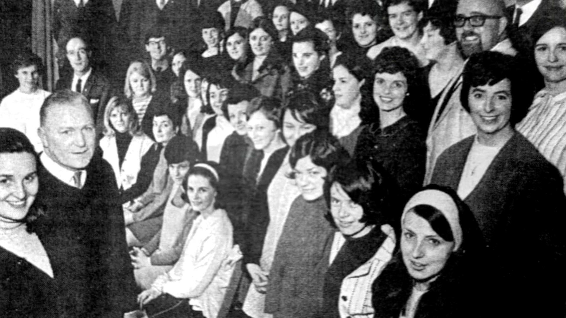 The cast of the first ever production staged at Edendork Hall in June 1963 - a group of mostly women in a black and white image with a few men. Some are wearing a nuns habit others in white/black tunics. All the men are wearing suits and everyone is smiling at the camera. 