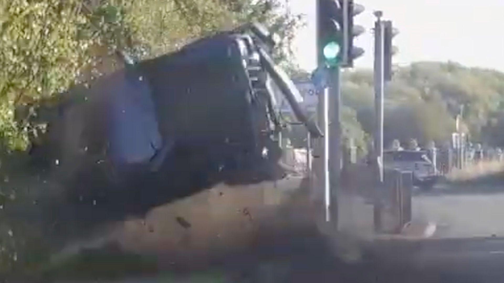 A car rolls on its side in the air at the side of a road, just before a set of traffic lights and a roundabout in the background.