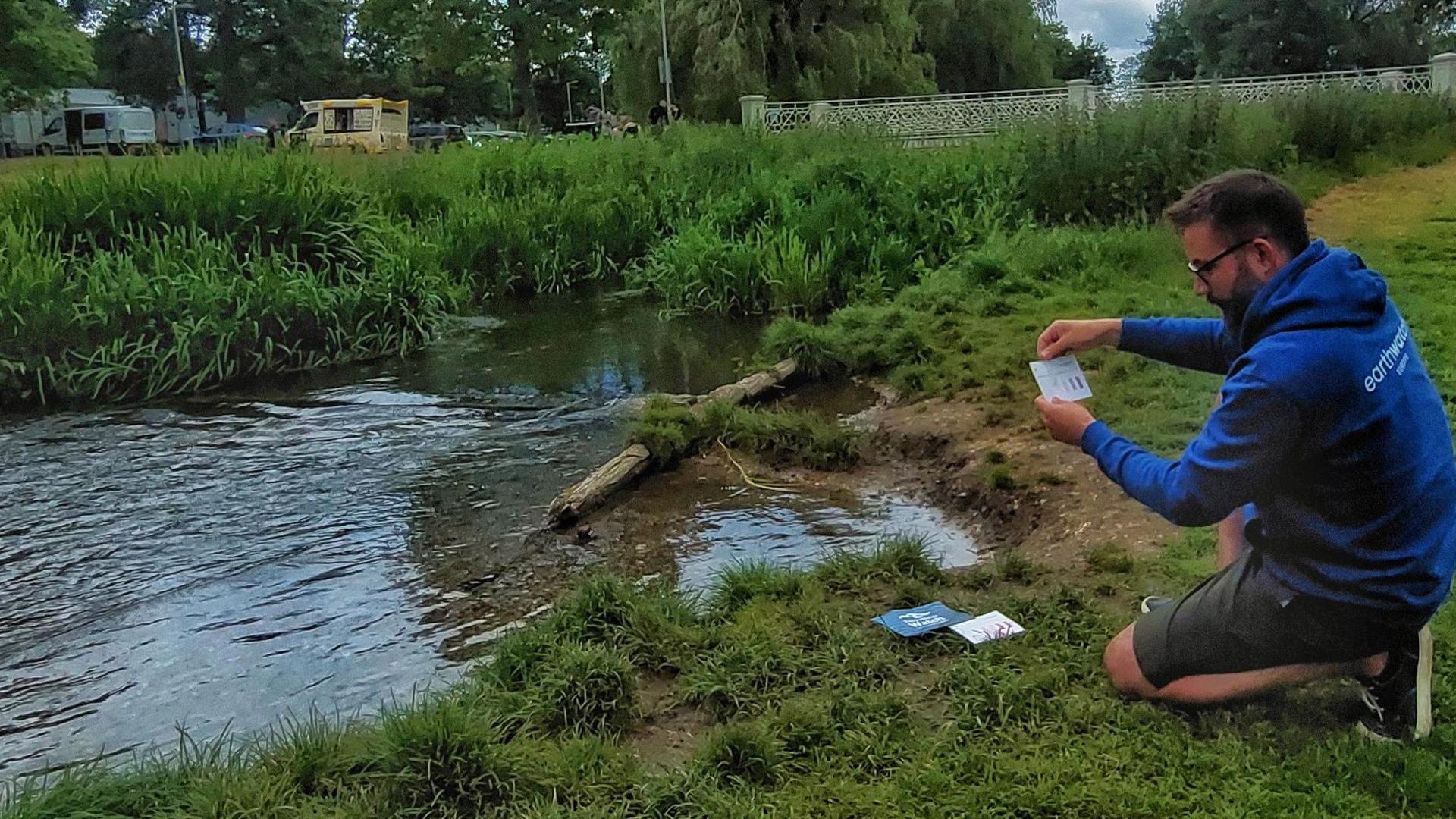 A small river flowing next to a park and a man kneeling next to it holding a piece of card with PH samples on. He is wearing a blue hoodie and shorts.