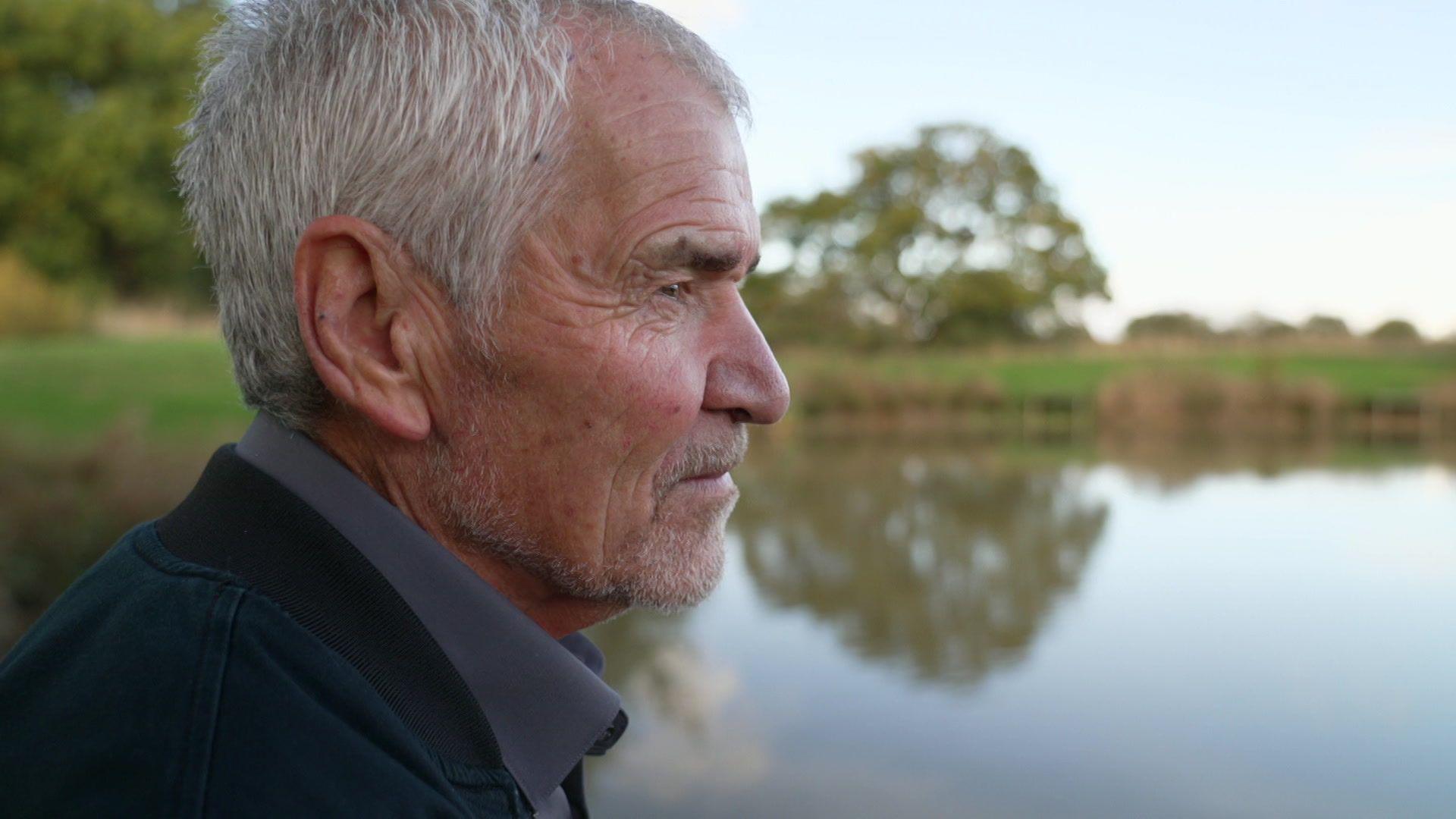 A close-up photograph of Dave Wyness as he looks out over a lake. He has grey hair and a short grey beard. He is wearing a dark grey shirt and a navy blue jacket.