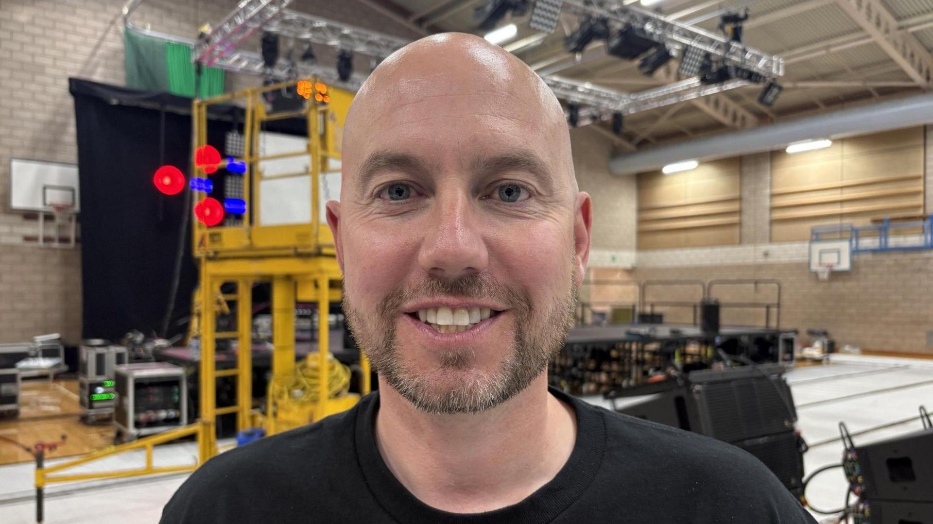 A man grinning at the camera. He is bald and is wearing a black top. He's standing in front of scaffolding and musical equipment in a sports hall. 