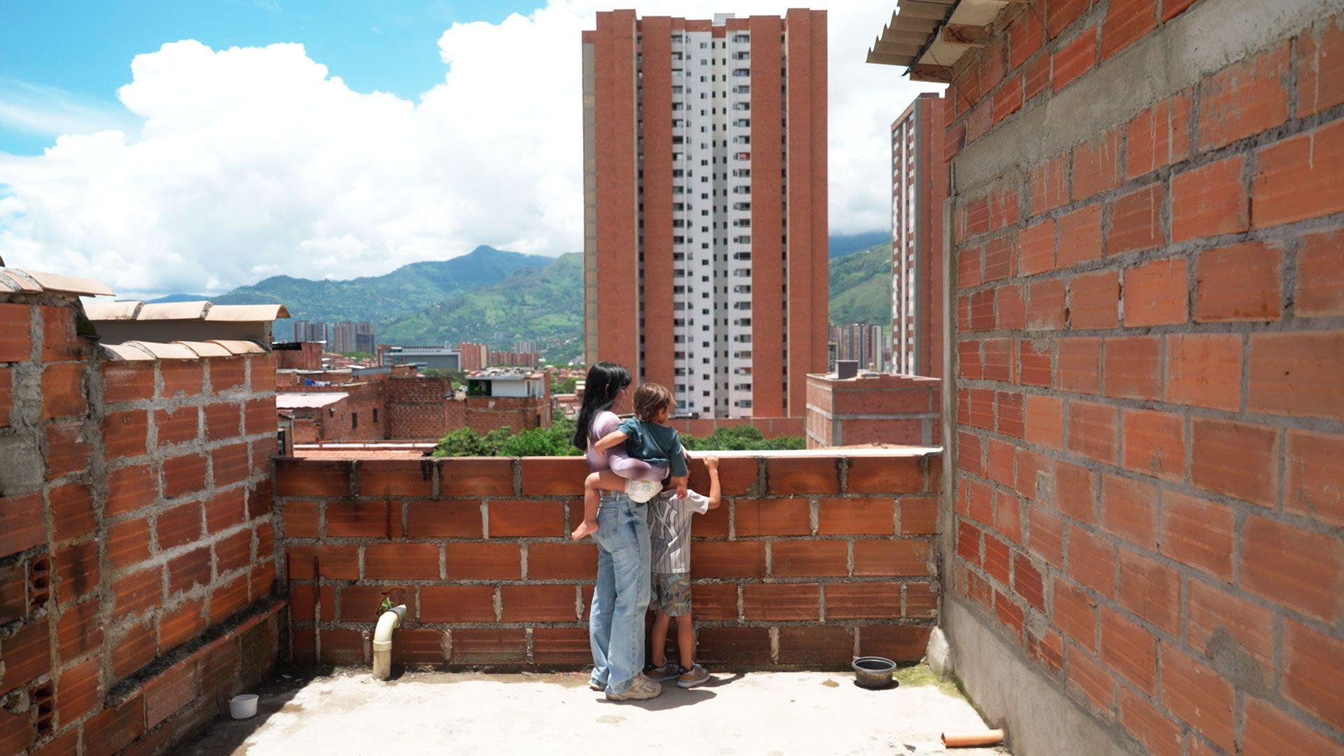 A young woman holding a toddler stands next to an older boy of about six or seven on a terrace looking out at a tower block and mountains in the distance.