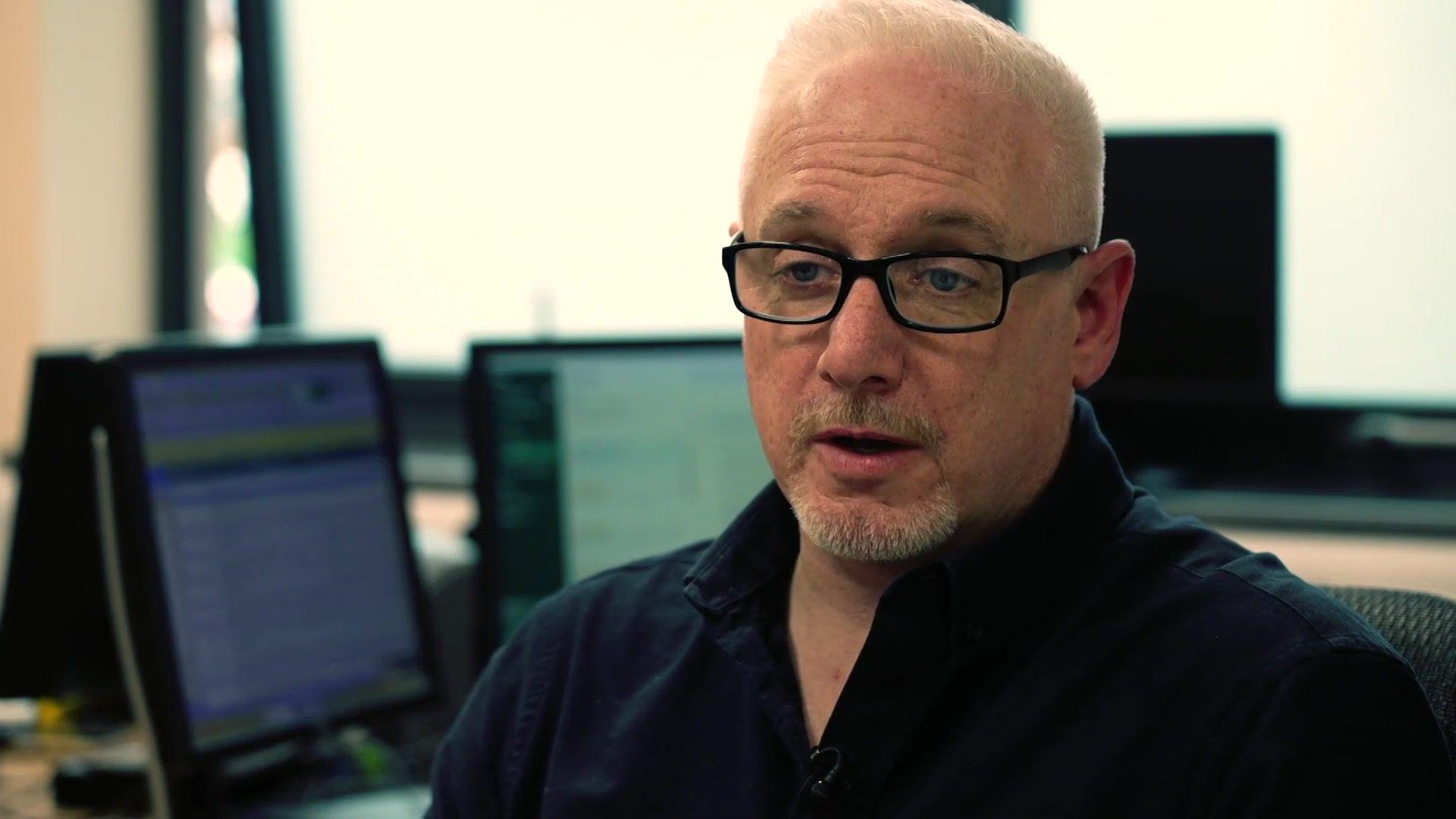 Dr Chris Scales has short grey hair and a beard. He is wearing dark framed glasses and a black shirt and is sat at a desk with two computer monitors in the background.