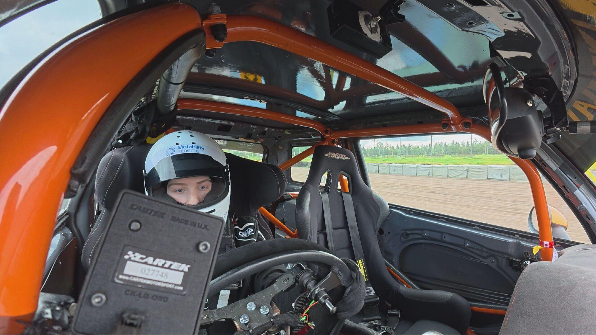 Ally Chalmers sitting at the wheel of a racing car. He is wearing a white helmet. The photograph is taken from a camera mounted on the car's dashboard