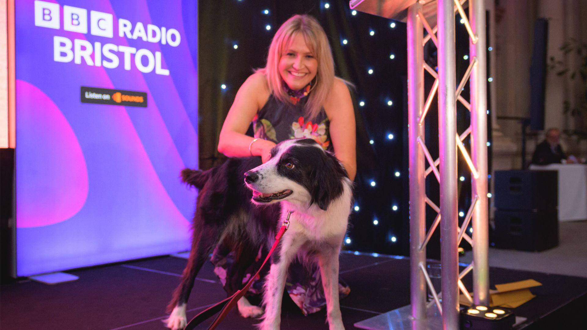 A black and white border collie cross stands in the centre of the stage whilst looking left in front of Claire Carter. She is blonde and wearing a black halter neck dress and is crouched down behind the dog.
