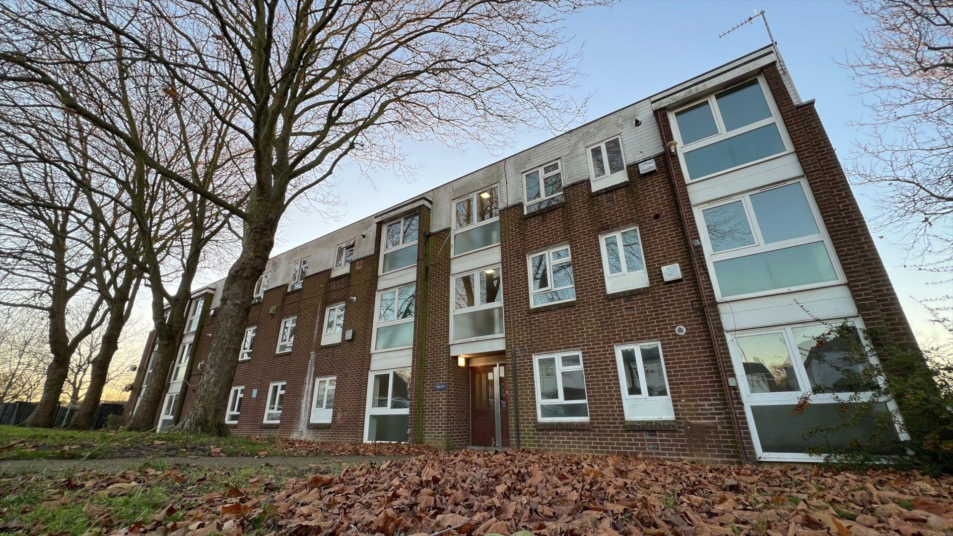 Sycamore Field flats in Harlow, with autumn leaves on the ground. There are a line of four trees. The block of flats is three storeys high. There are blue skies above.