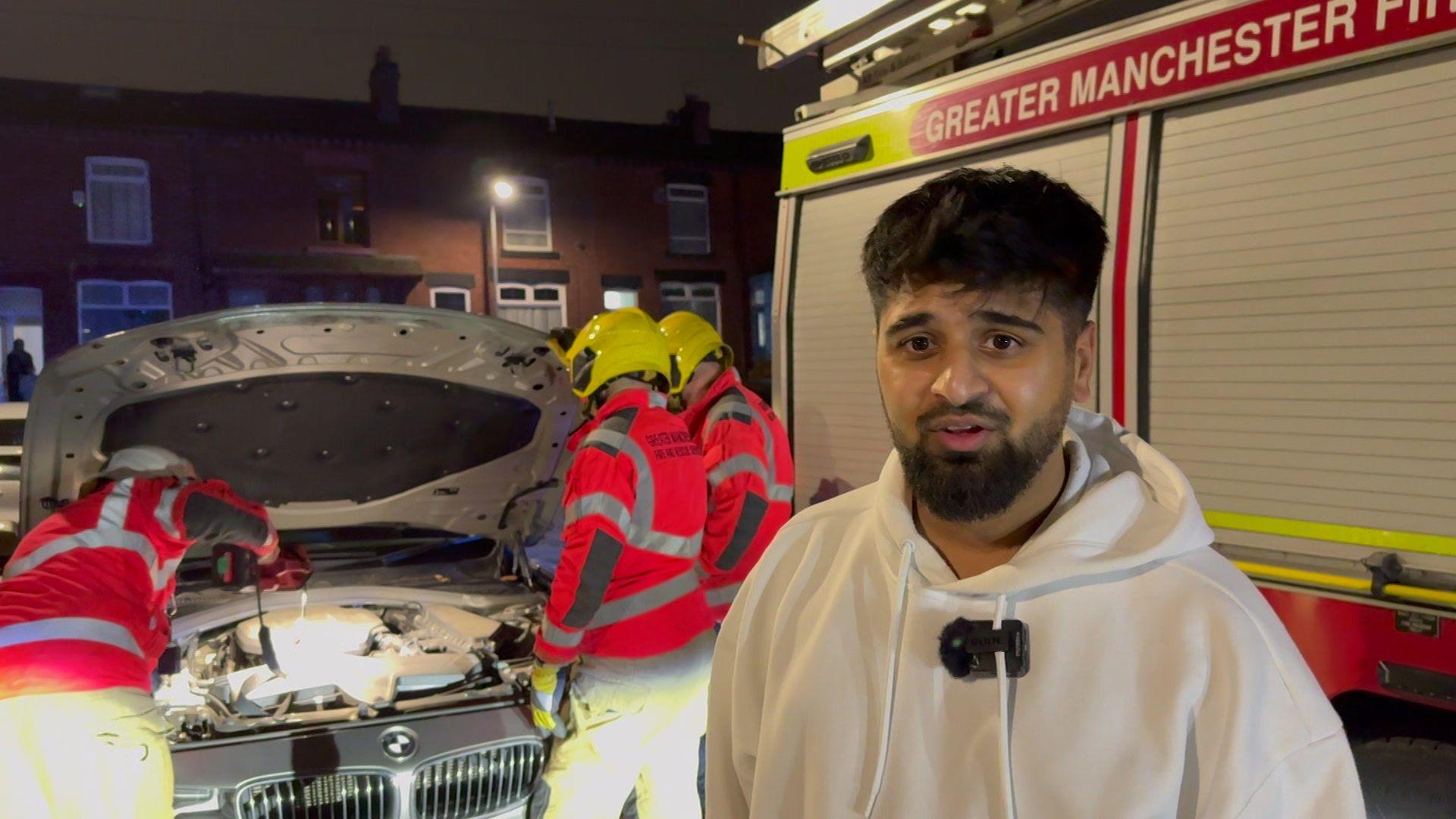 Nizam Pate stands in front of his grey BMW. The bonnet is up and the firefighters are stood over it while. Nizam has short dark hair, he wears a white hoodie. A fire engine can be seen in the background.