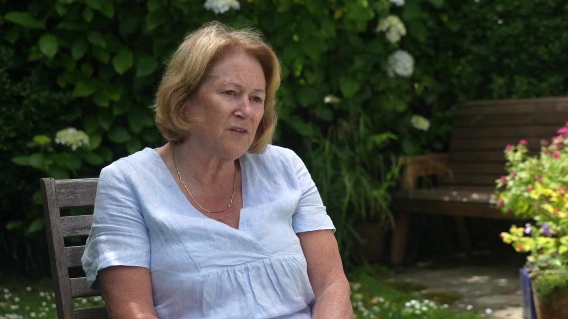 Linda Taylor sitting on a wooden chair in a garden, wearing a light blue top 