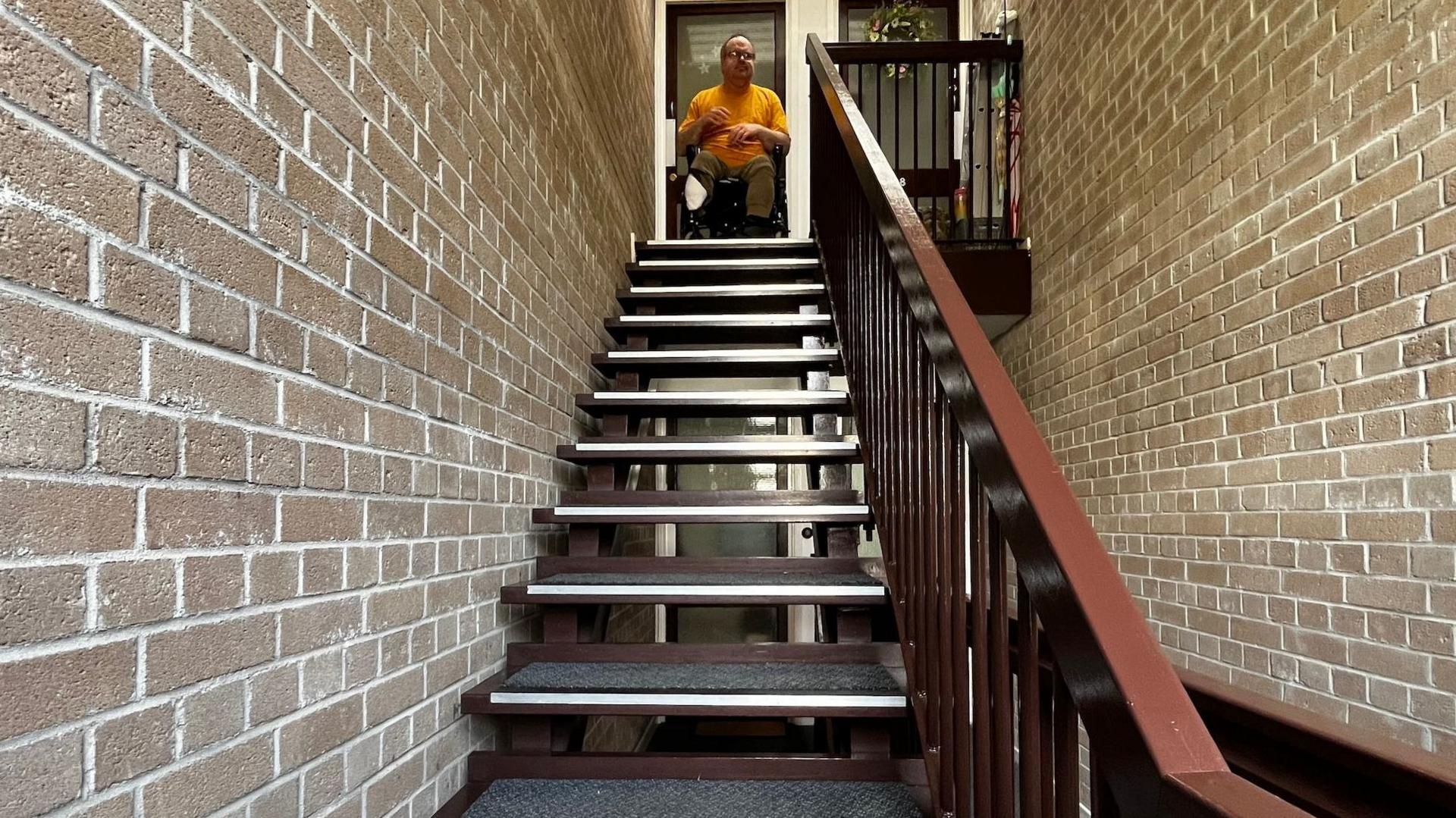 James Smith, a man in a wheelchair, is sitting in the open front door of his flat at the top of a flight of stairs. The stairs are open stairs - so there is space between each one - and a wooden handrail is to the right. 