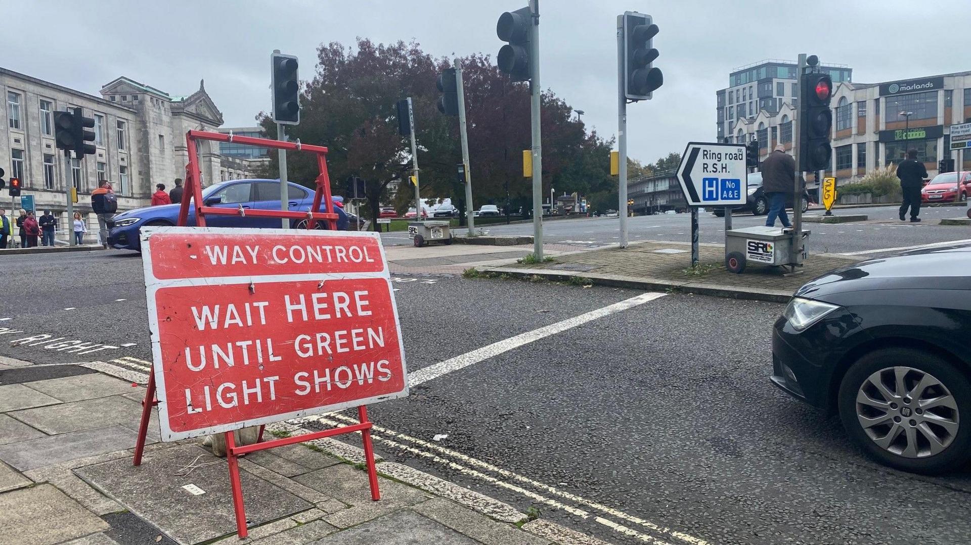 Red temporary traffic sign at junction which reads: "Wait here until green light shows"