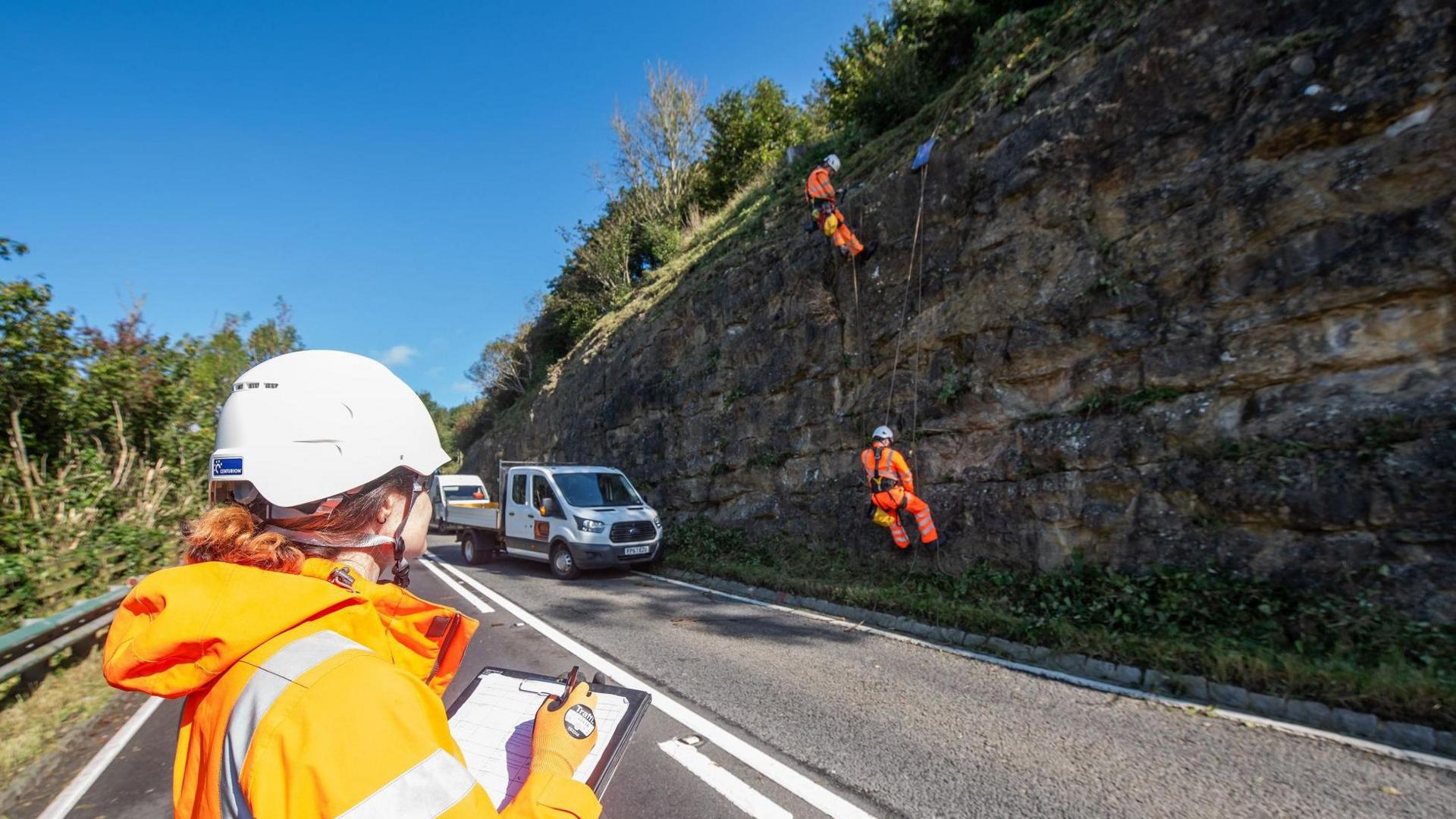 A person in a high vis jacket holding a clipboard and wearing a white helmet looks at a rock face. On the rock face are two people in full orange high vis abseiling. On the road below is a parked white van.