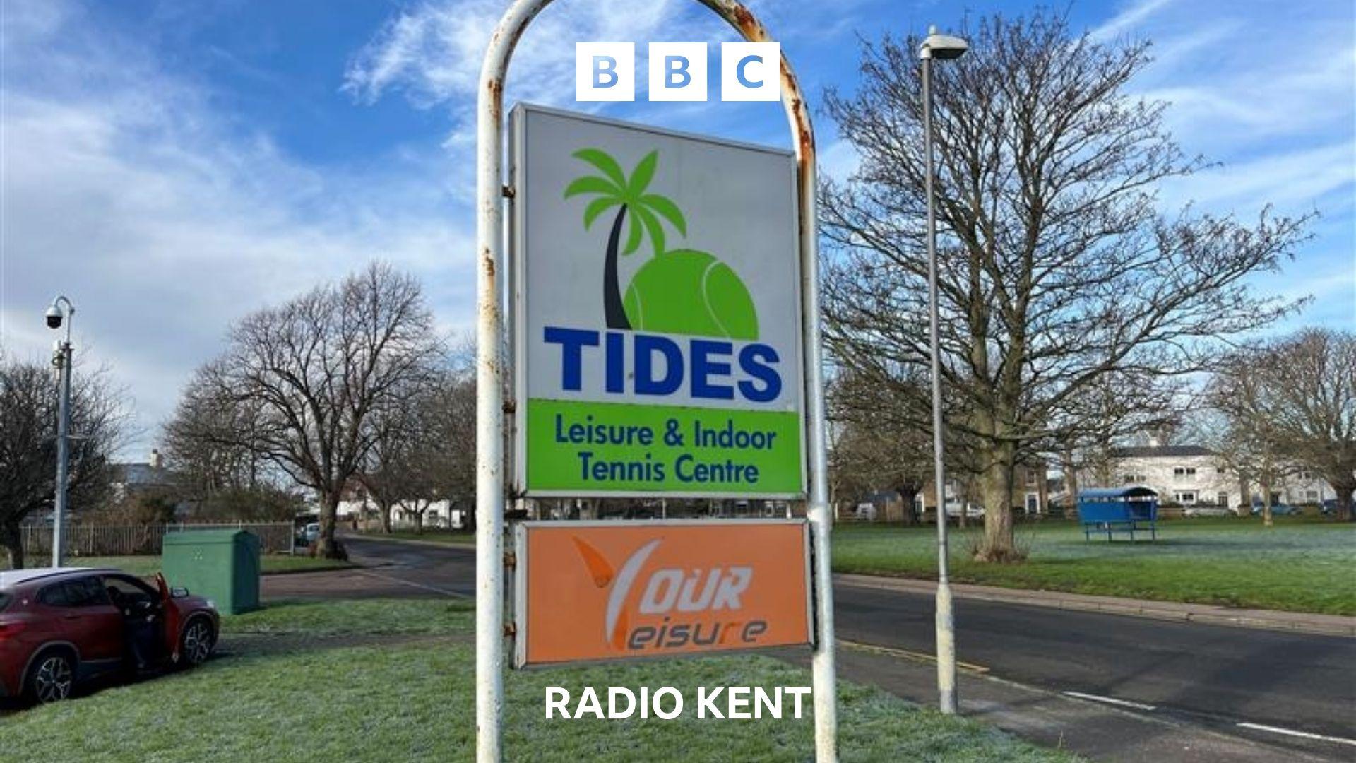 The white green and blue 'Tides' sign is shown outside the entrance to the leisure centre. The logo depicts a green palm tree and tennis ball.  A residential area is behind the sign with grassy areas, a road and trees. There are houses in the distance.