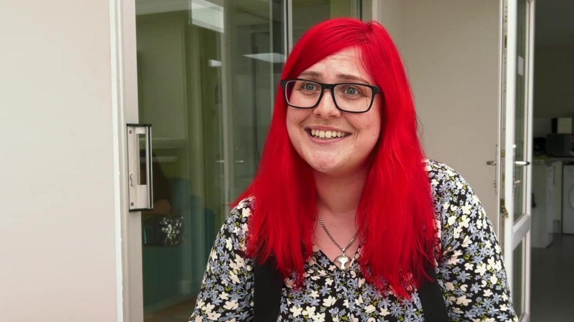 Laura Watts, who has long red hair and is wearing glasses and a flowery blouse, smiling at the camera. There is a white wall and a glass door behind her