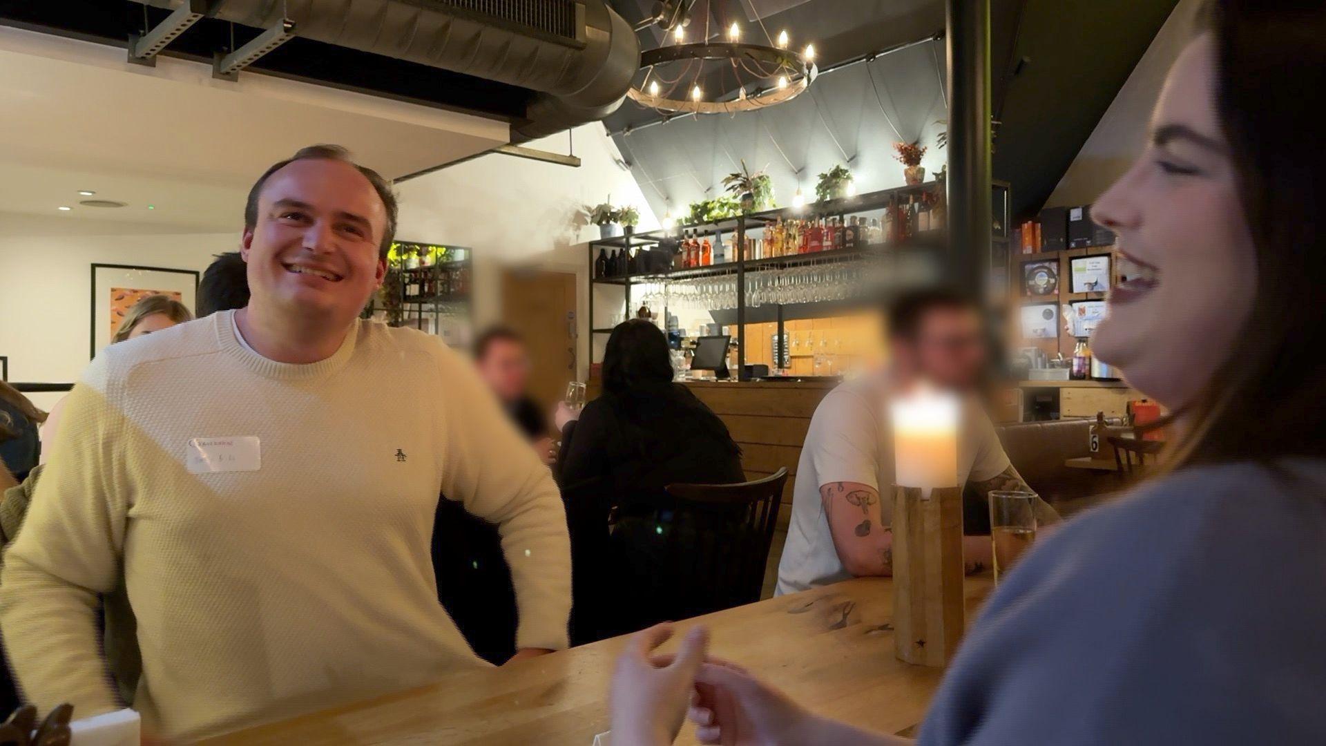 A man and woman are sitting across from each other at a speed dating event. The man is wearing a white jumper with a name badge on. The woman is wearing blue. They are laughing between one another. 