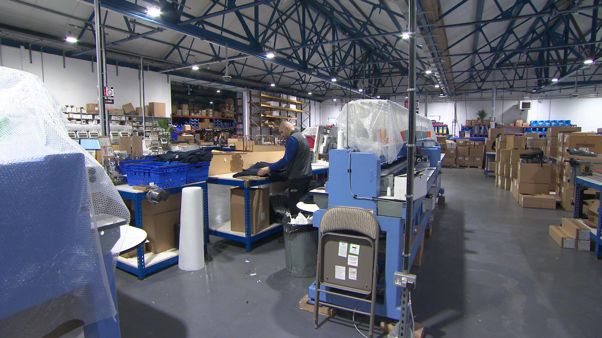 Man folding a t-shirt at a warehouse next to a textile machine and stacks of brown cardboard boxes