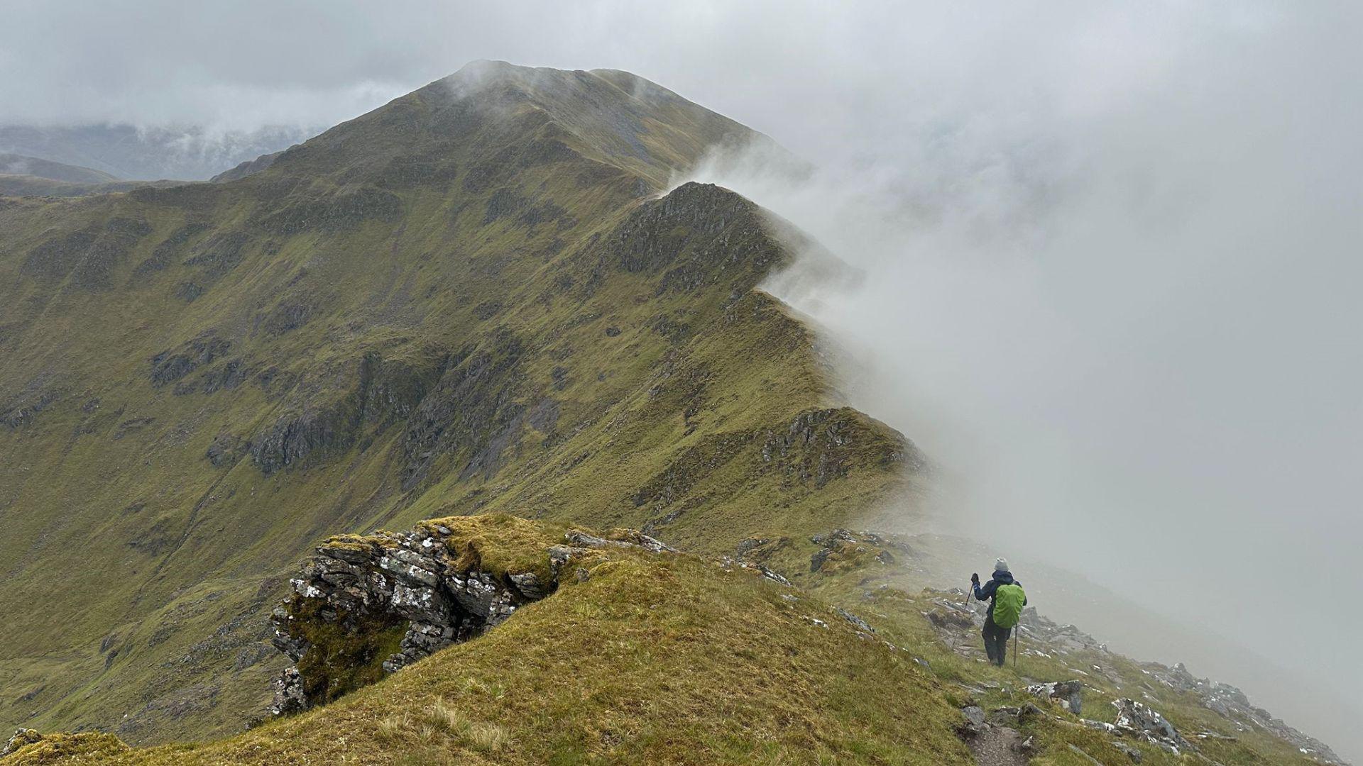 A mountain ridge covered in grass with a hiker wearing dark clothing and a light green backpack walking with poles in the foreground. Part of the left side of the image is covered with a thick white-grey fog. The sky is overcast.