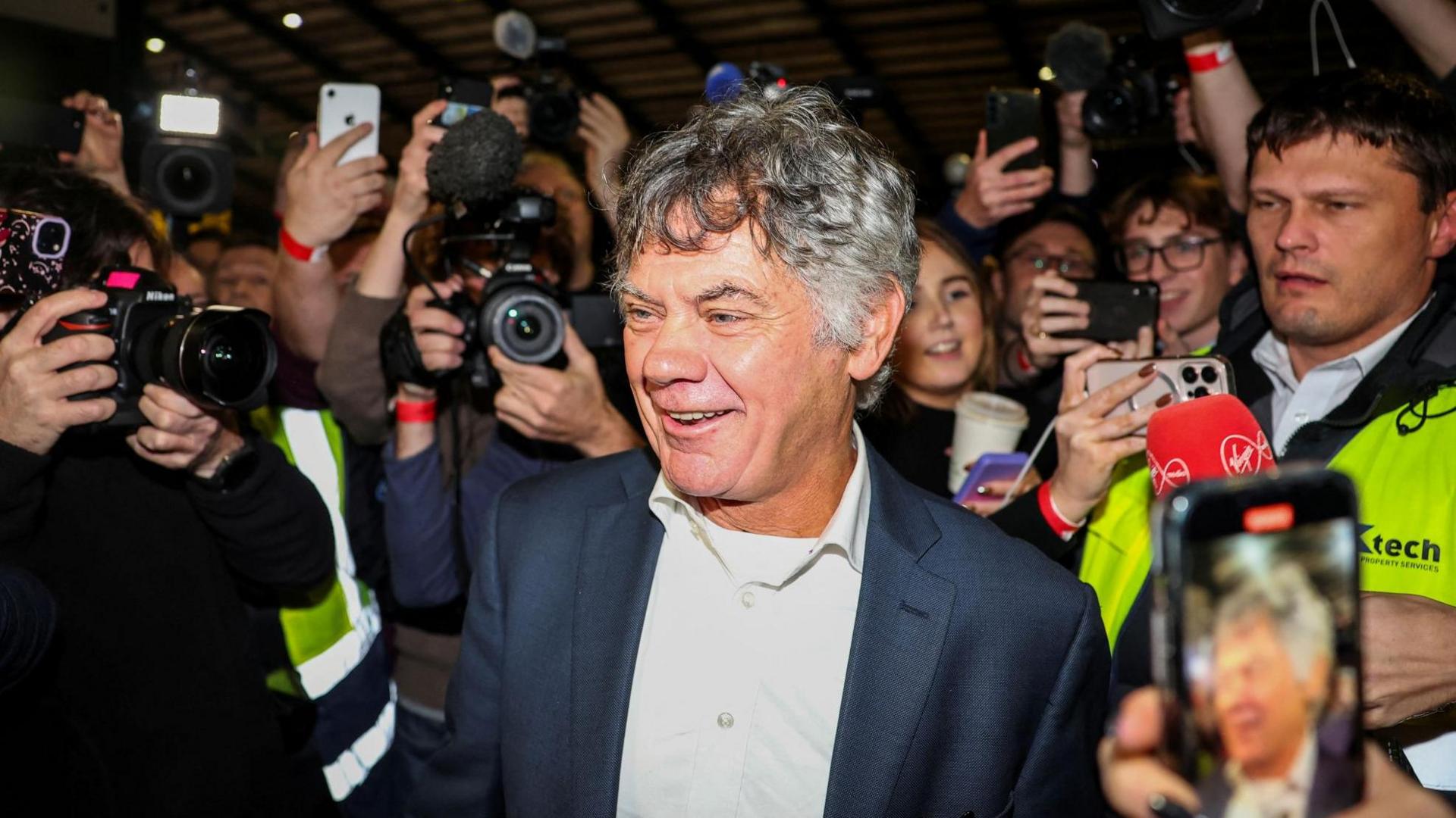 Gerry Hutch, who is an older man with dark, greying hair is surrounded by a media scrum as he enters a count centre in Dublin. He is wearing a dark jacket and white collared shirt.