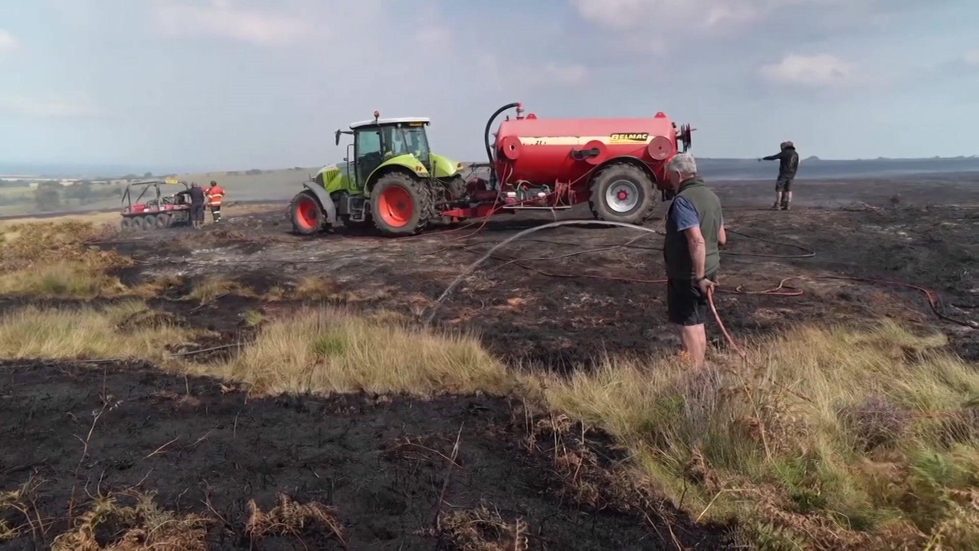A man sprays water from a hose onto charred moorland, in front of a green tractor and red tanker, while other men also spray the ground with water from hoses