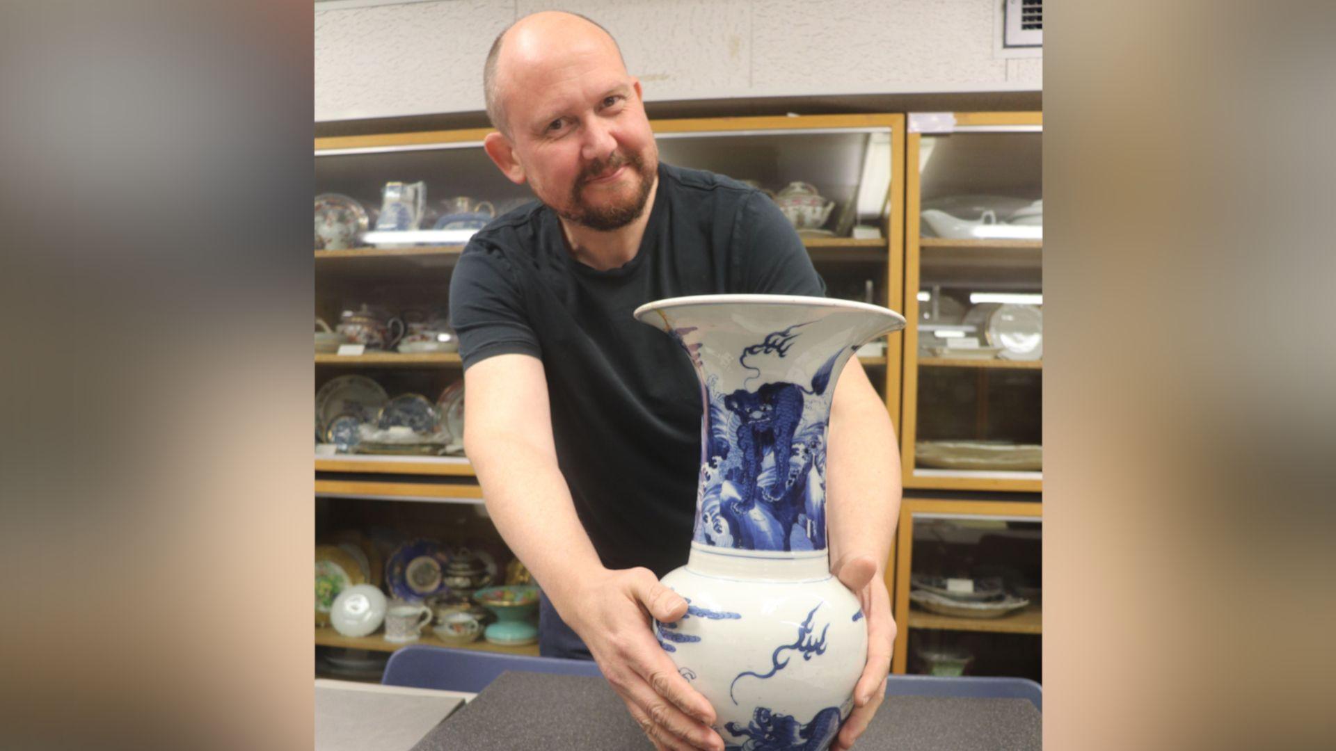 Professor of ceramics Neil Brownsword, holding a blue and white vase, is wearing a black t-shirt and is standing in front of a glass cabinet with a number of ceramic items in it. He has cropped brown hair and a trimmed beard and moustache.