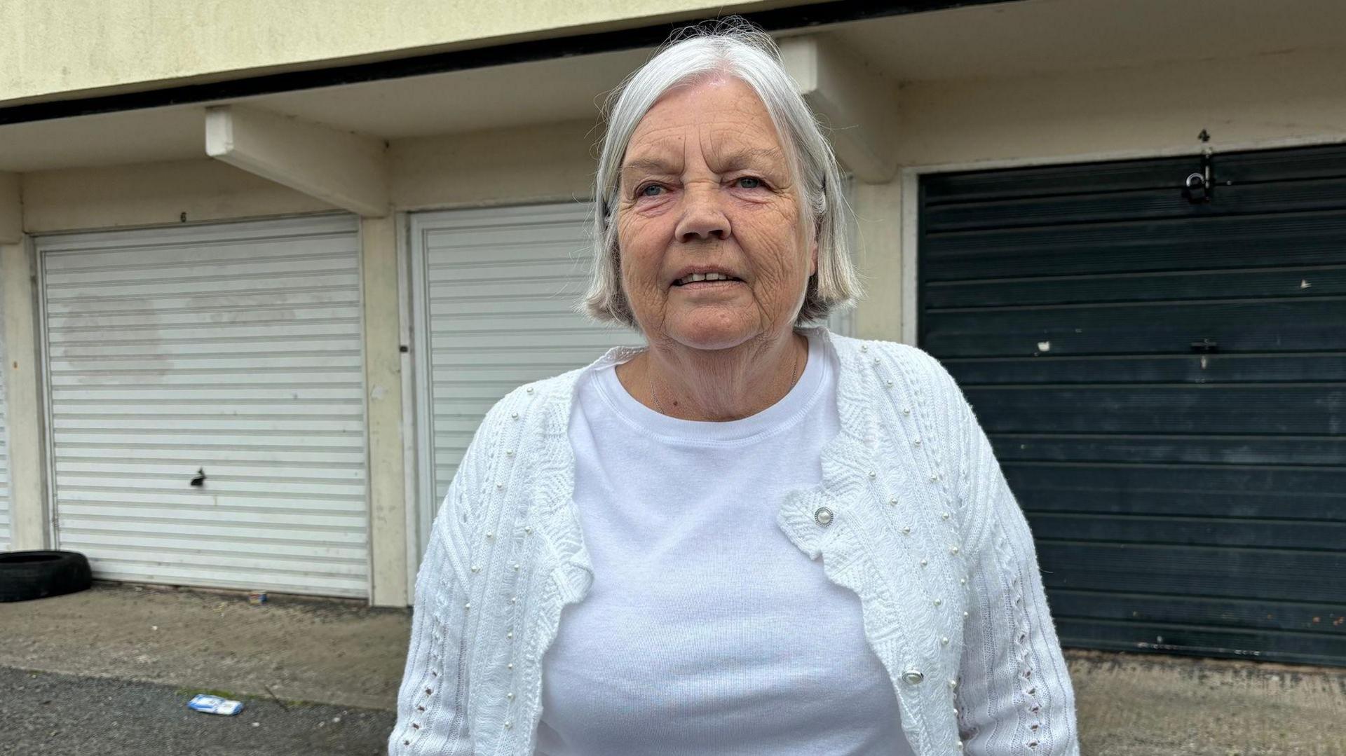 A woman with grey hair, wearing a white top and cardigan, stands in front of a row of closed garages.