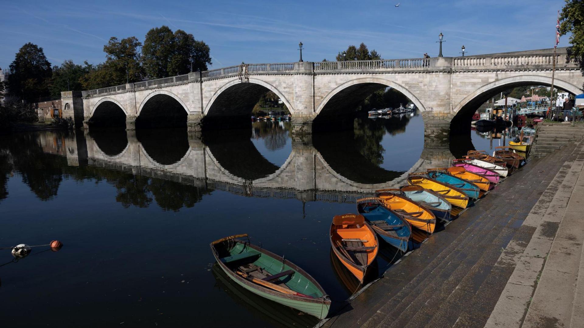 Library image of the Thames at Richmond, on a sunny day. Richmond Bridge is the main focal point of the photograph, with small rowing boats of various bright colours tethered to the concrete steps leading down to the water.