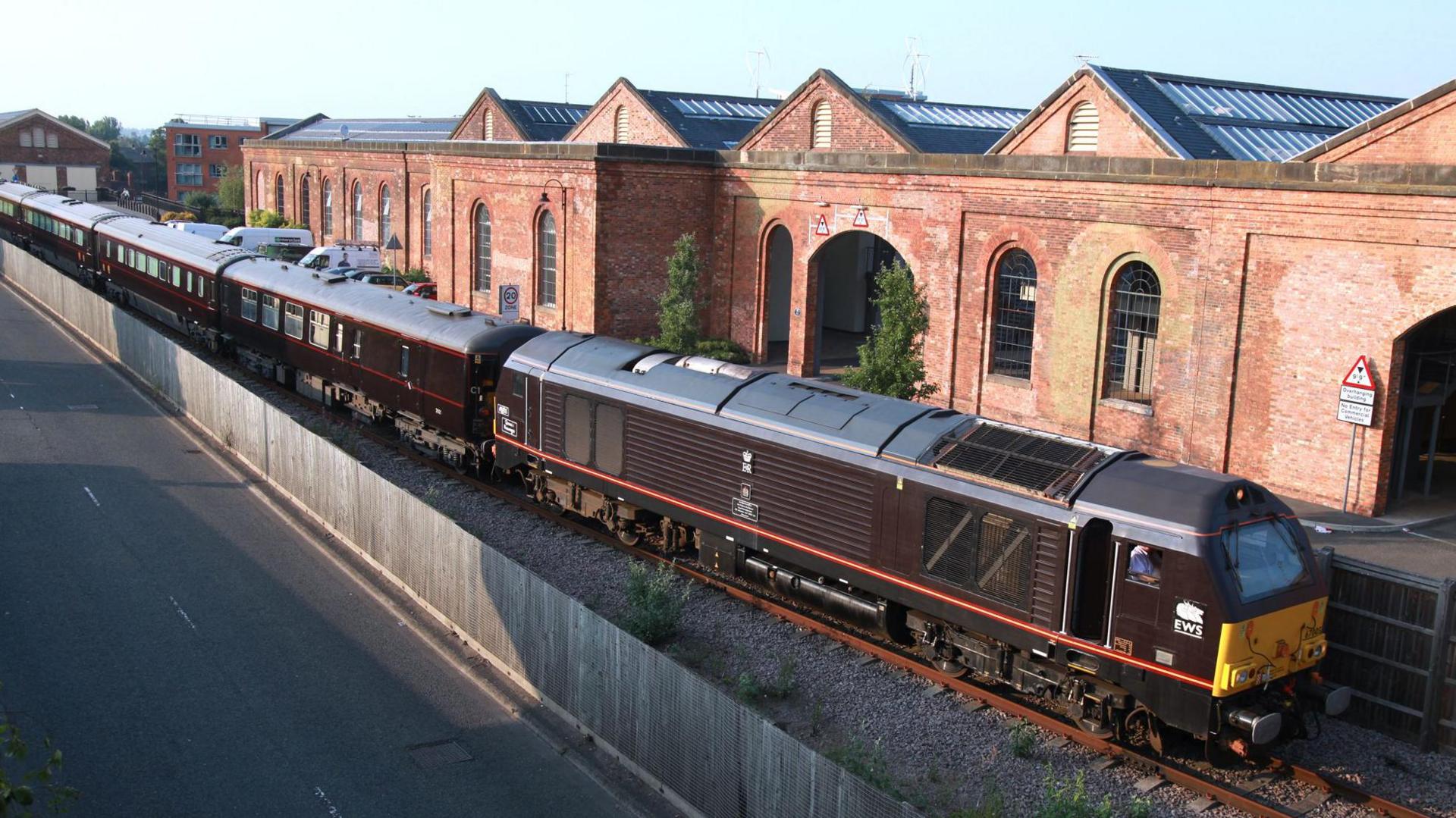 Five carriages of the royal train are visible on a track, behind it is the old red-brick buildings of Wolverton Works