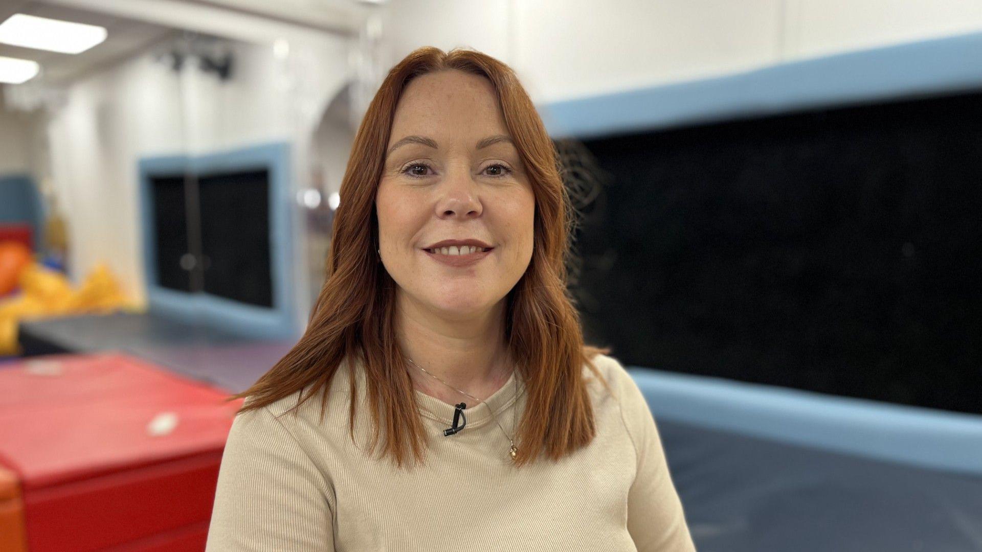 A woman smiles and looks directly at the camera. She has auburn, mid-length hair. She is wearing a cream crew-neck, long-sleeved top. Although the background is blurred, it is colourful - a children's soft-play area.