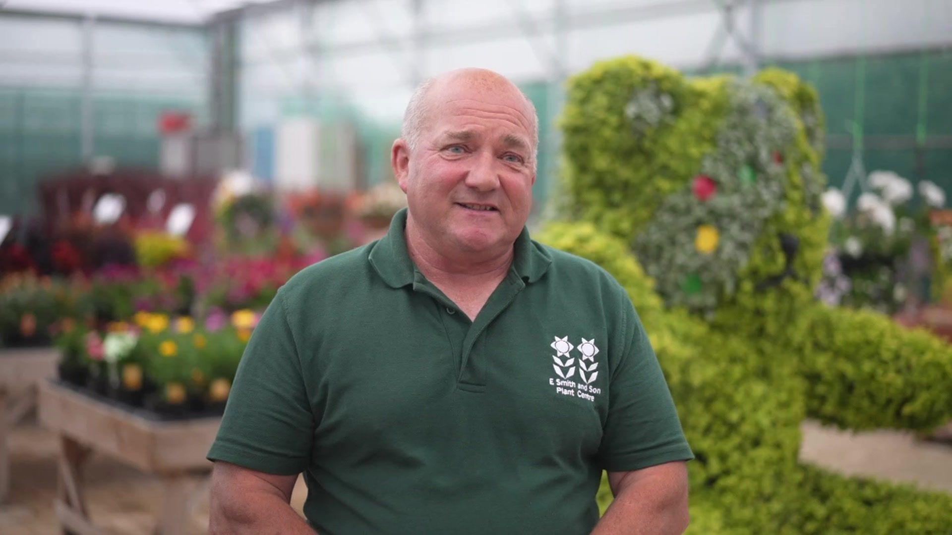 Derek Smith standing in front of a giant Pudsey bear made from flowers planted in a metal frame. Derek is wearing a green polo shirt with two flowers on the emblem and it reads E Smith and Son Plant Centre