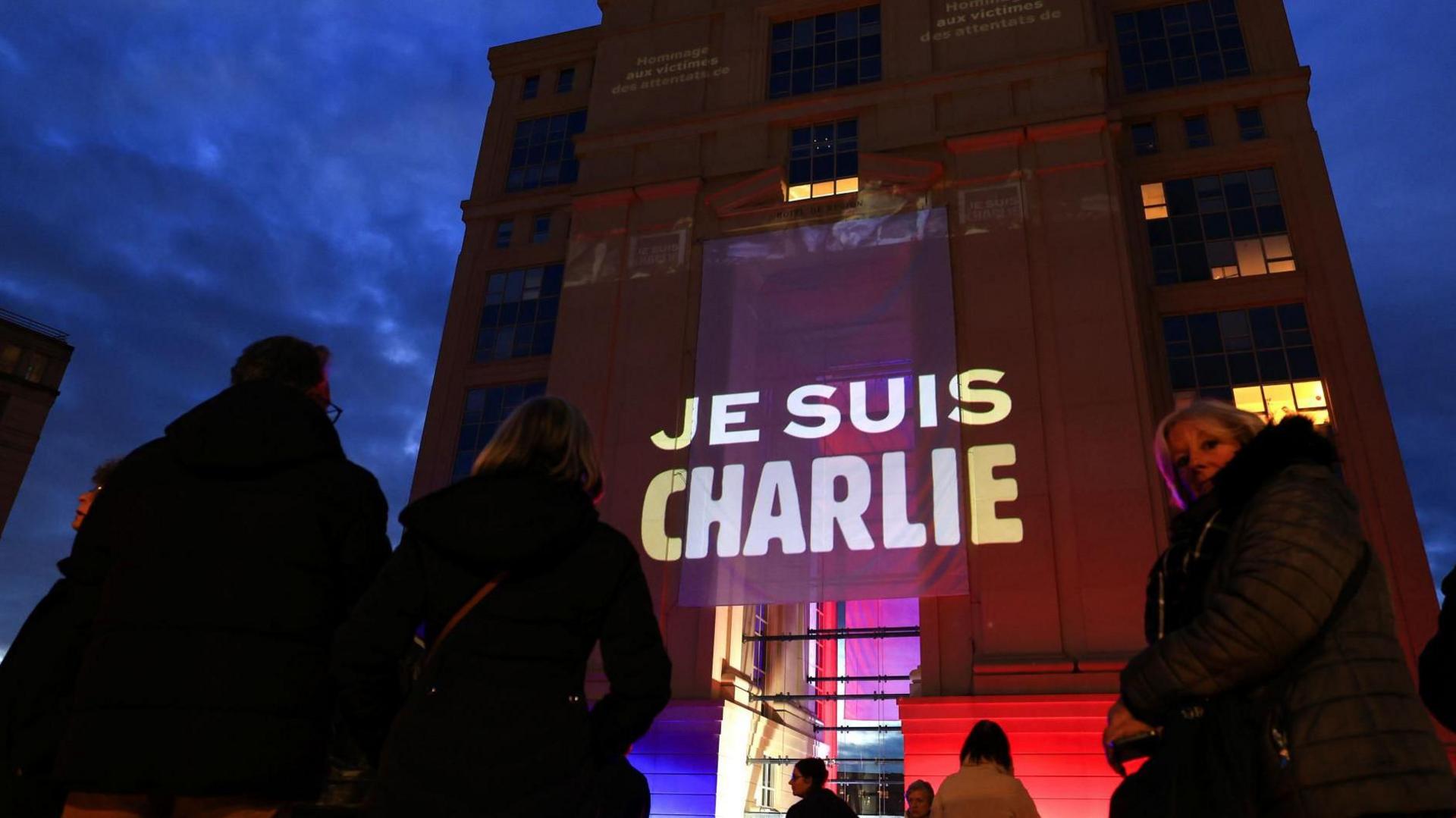 People stand outside a building draped with a banner stating "Je Suis Charlie" or "we are Charlie"