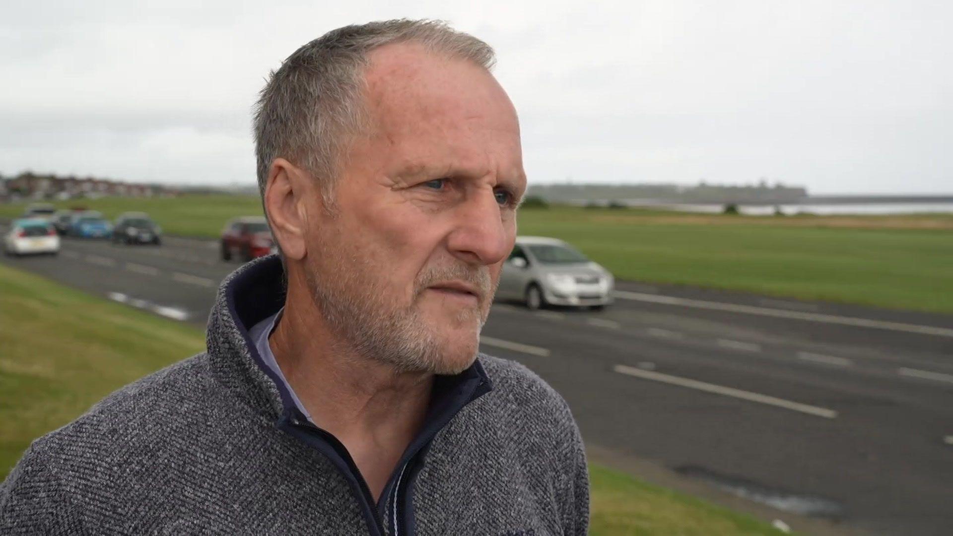 Dave Dixon, campaigner, with short grey hair, wearing a grey fleece, looks to the right as he stands on a grass field, with a road with cars driving past next to him. 