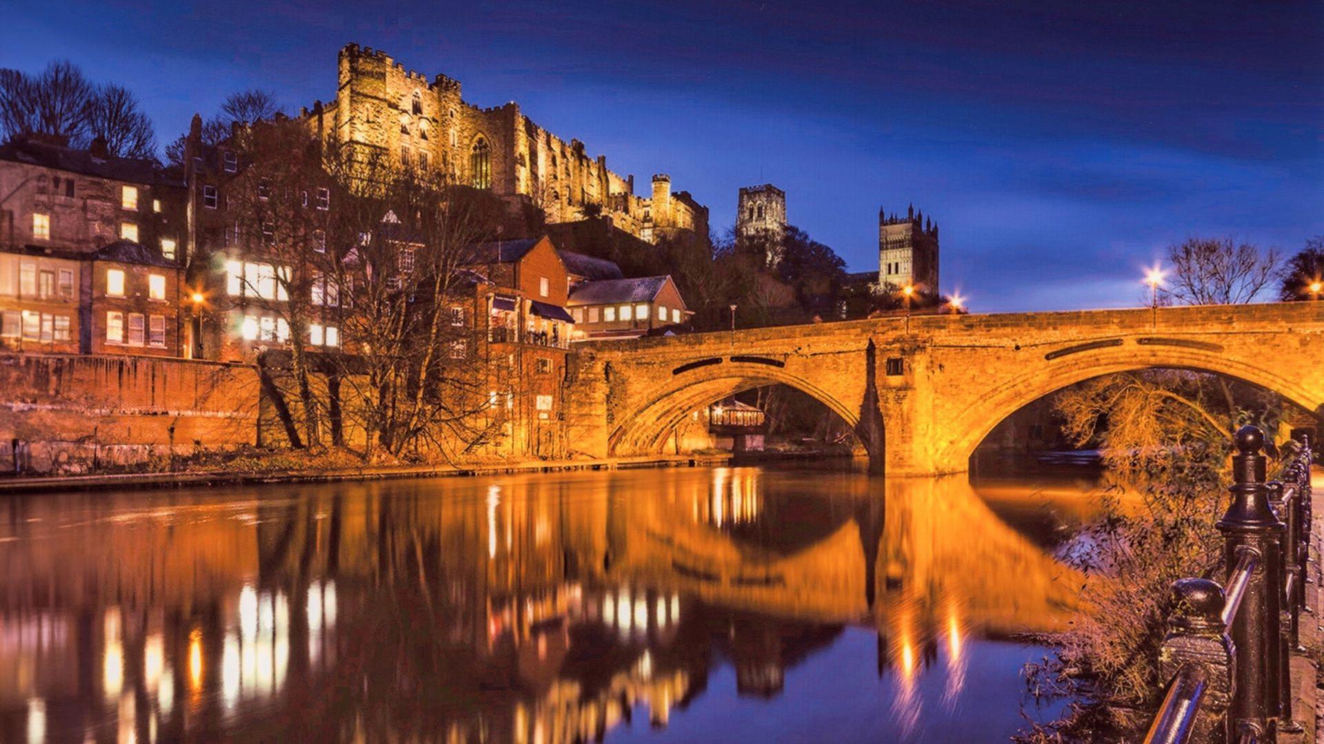 An old bridge with two arches cross a river, with a castle and cathedral beyond it lit up at night, all reflected in the river.