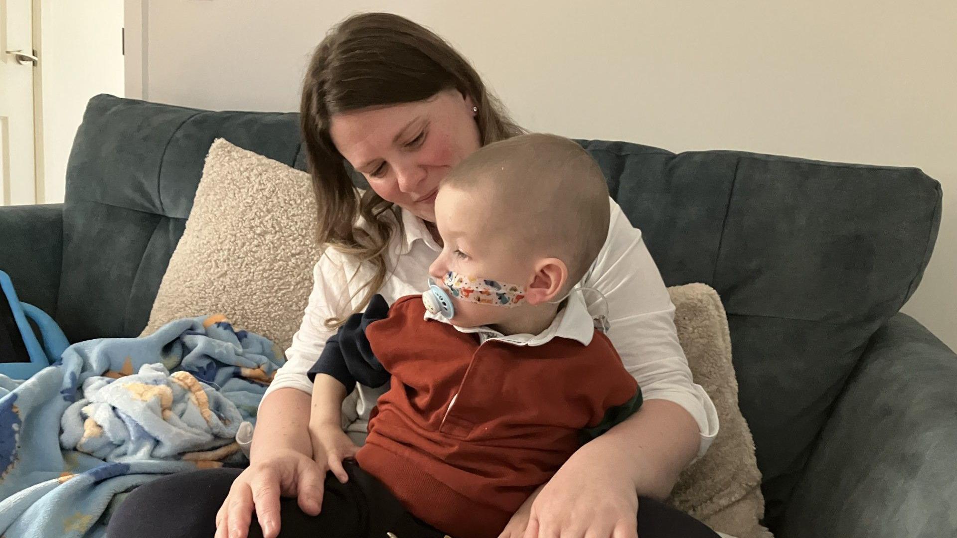 The photo shows Deborah Rowe with her son Jonah sitting on her lap. They are sitting on a grey sofa, with a cream wall behind them. There are cream cushions behind Deborah. Deborah has long, dark brown hair and she is looking lovingly at Jonah. He is looking to the left. He has a dummy in his mouth, and a nasogastric tube stuck to his cheek with colourful tape and feeding into his nose. There is a toy aeroplane near his hand.