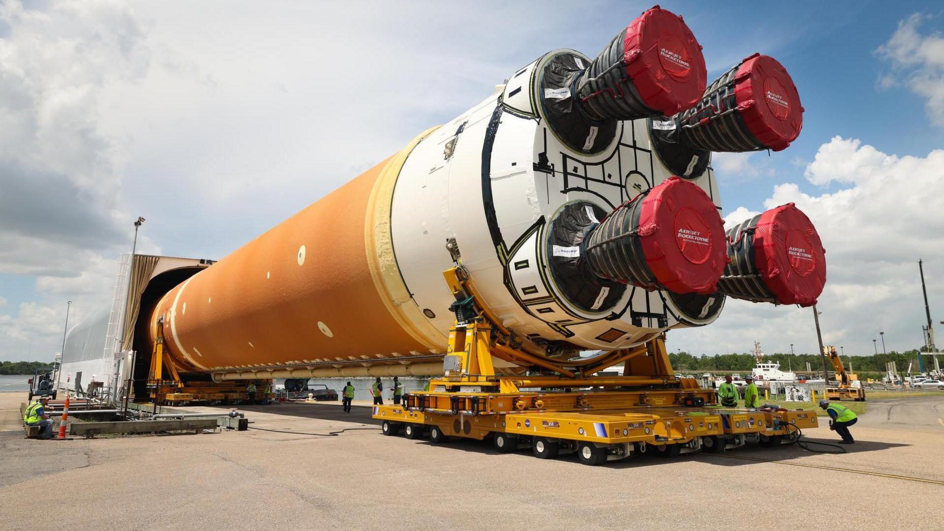 Nasa's SLS rocket lying horizontally and on supports and being wheeled into an enclosure. The rocket appears gigantic in compariosn to engineers sitting alongside the vehicle.