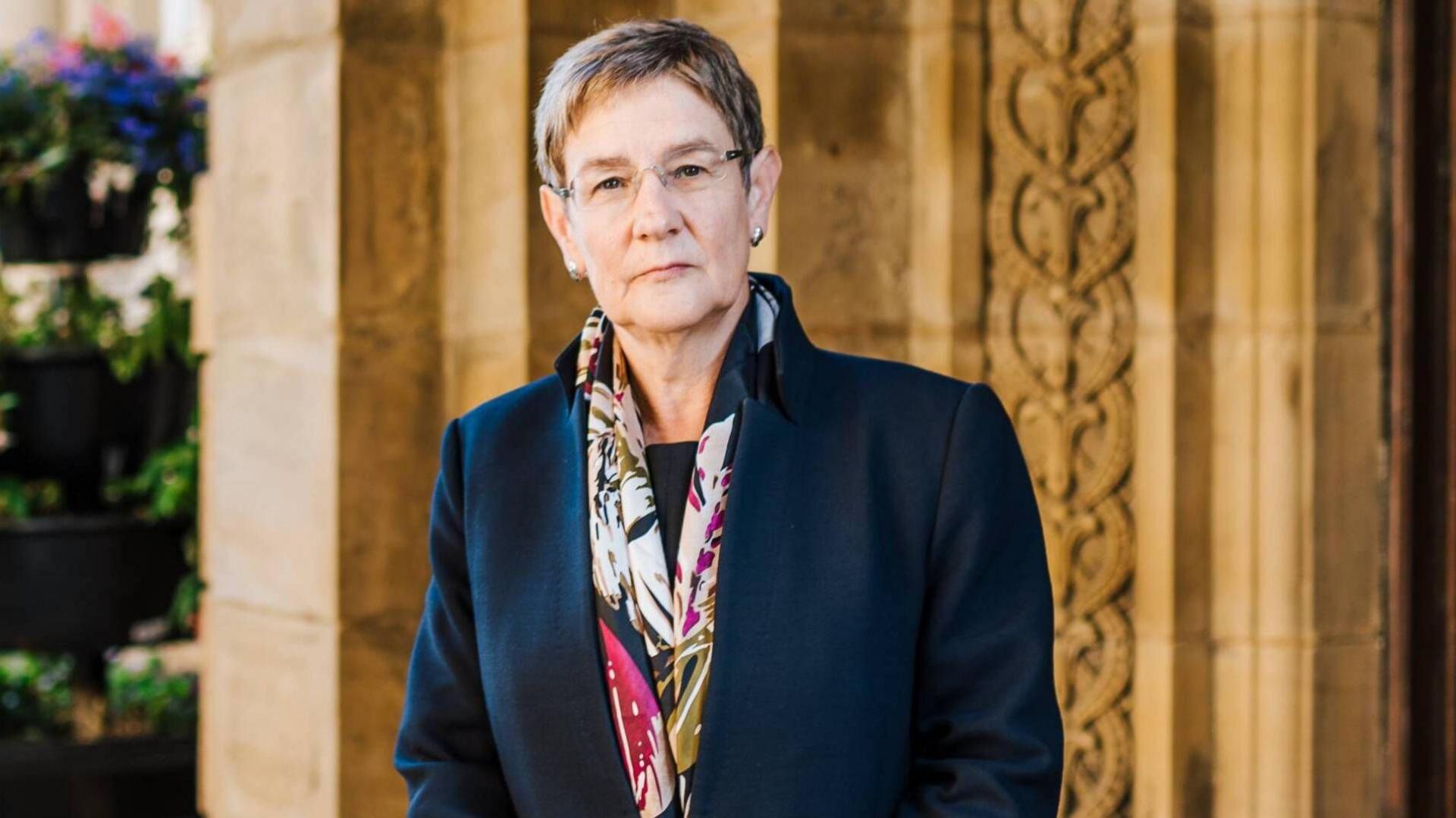 Woman with short grey hair and glasses wearing a blue coat with a colourful scarf